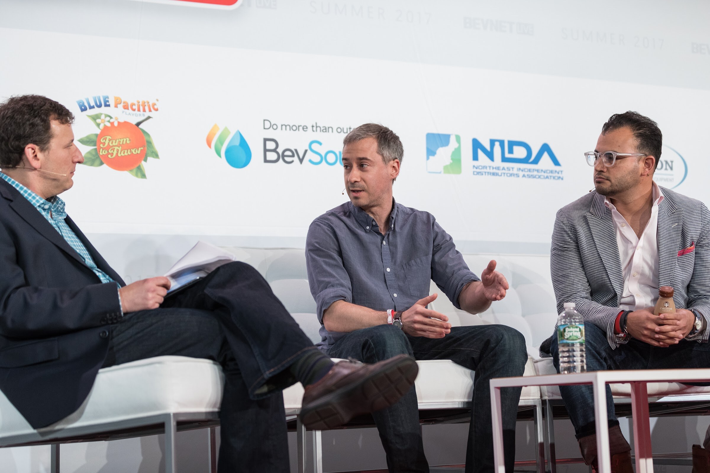 Three men seated on white chairs on stage; the one in the middle gestures while the others listen, with Blue Pacific Flavors, BevSource and NIDA logos behind them.