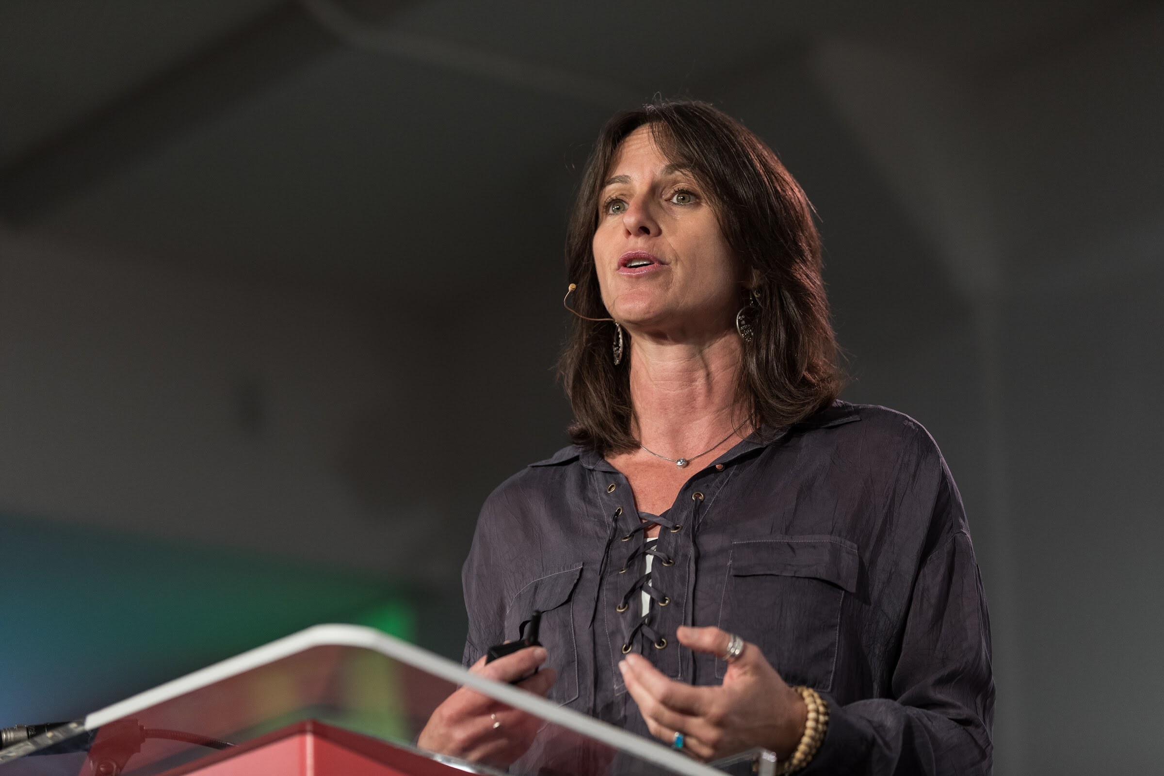 Woman with shoulder-length dark hair in a dark lace-up blouse speaking at a clear podium with a headset microphone.