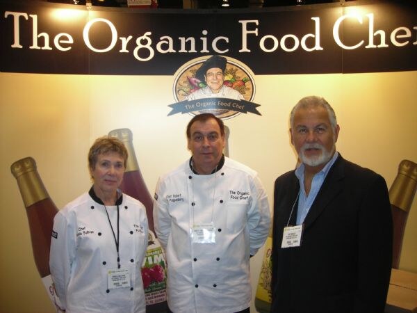 Two people in white chef coats and a man in a dark jacket stand together in front of a The Organic Food Chef sign and large bottle graphics.