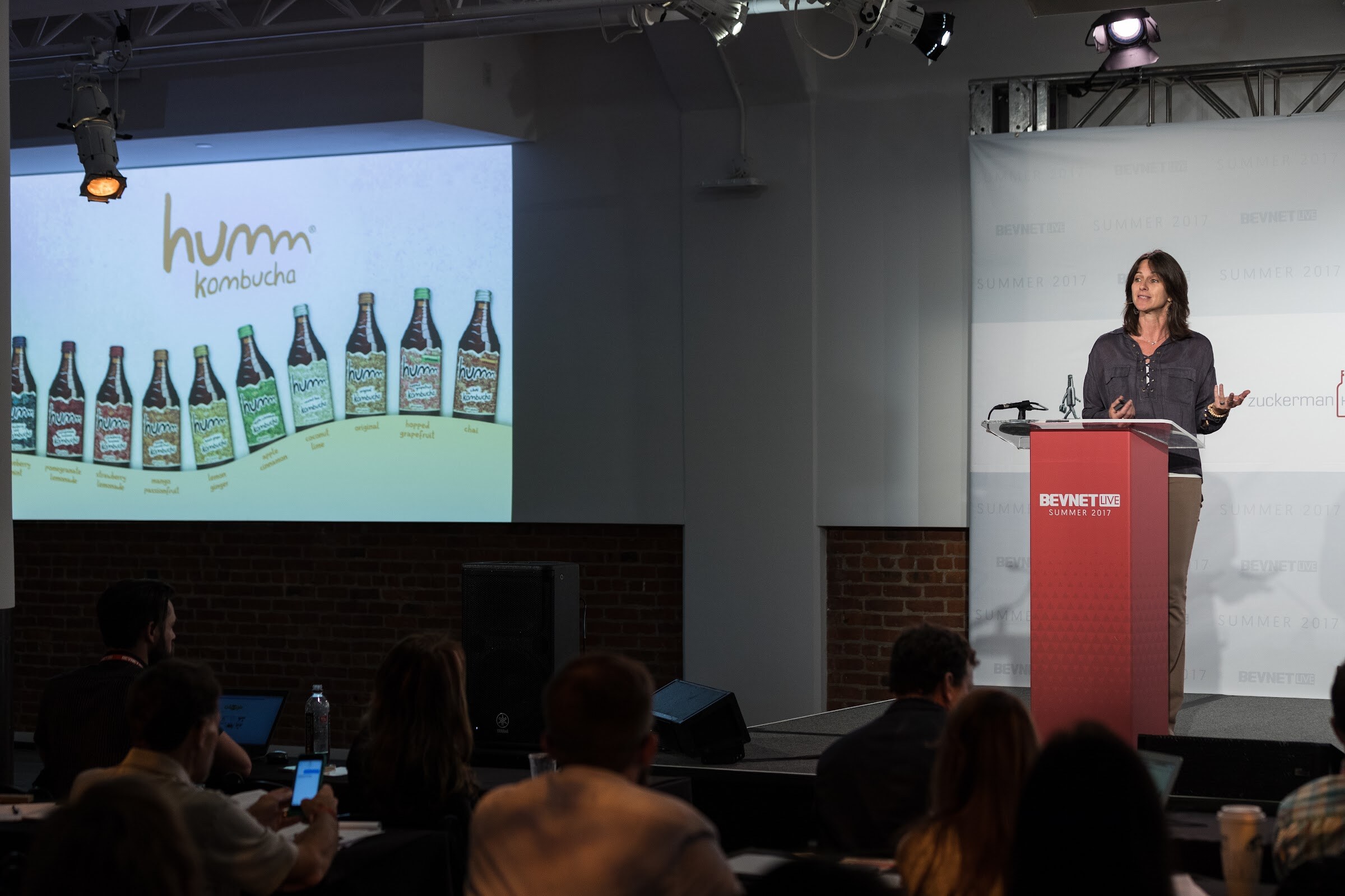 Wide view of a woman at a red BevNET Live Summer 2017 podium beside a projected slide reading humm kombucha above a row of bottles, with audience members in the foreground.