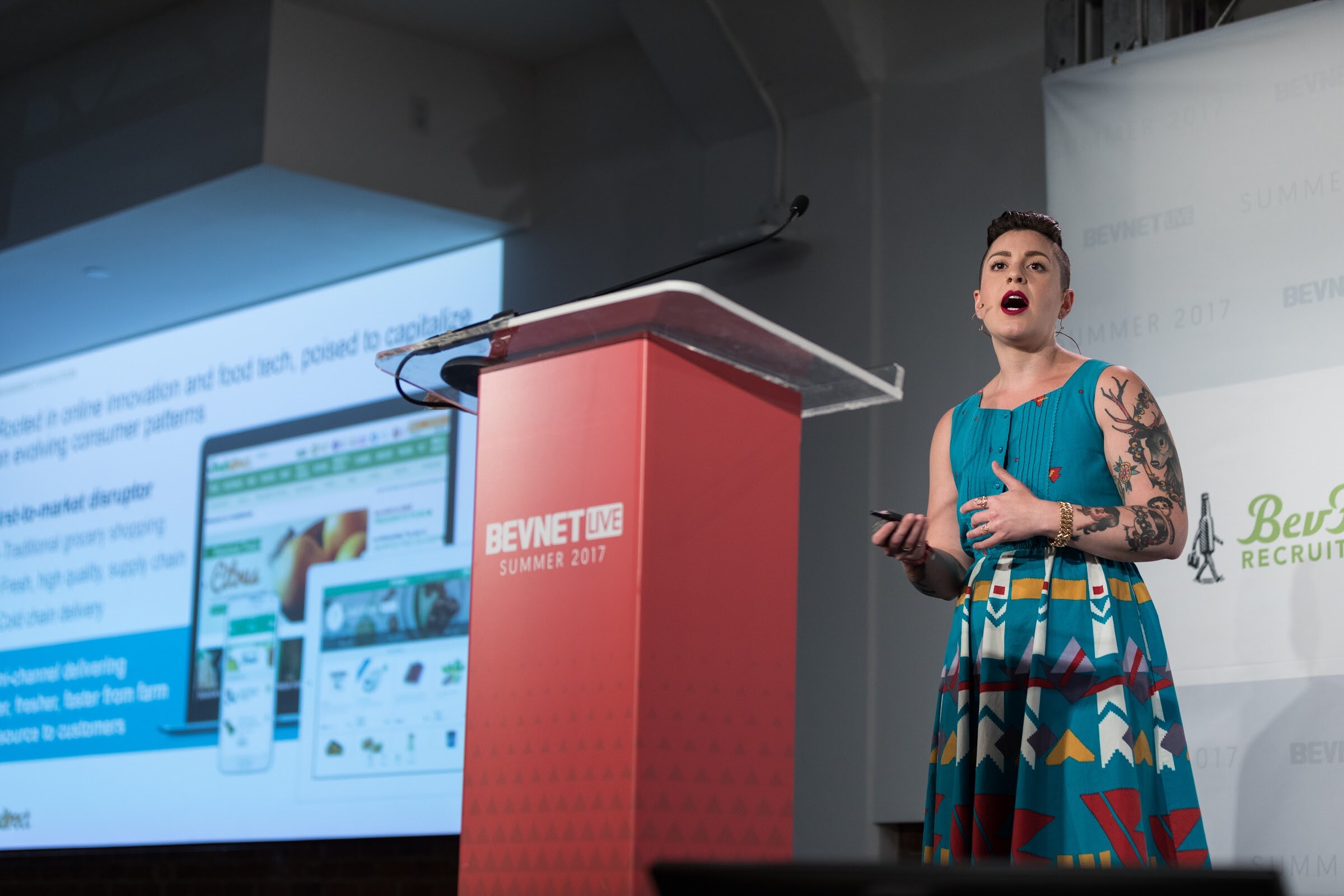 Woman in a teal patterned dress with tattooed arms standing beside a red BevNET Live Summer 2017 podium, with a projected slide about online food innovation to her left.