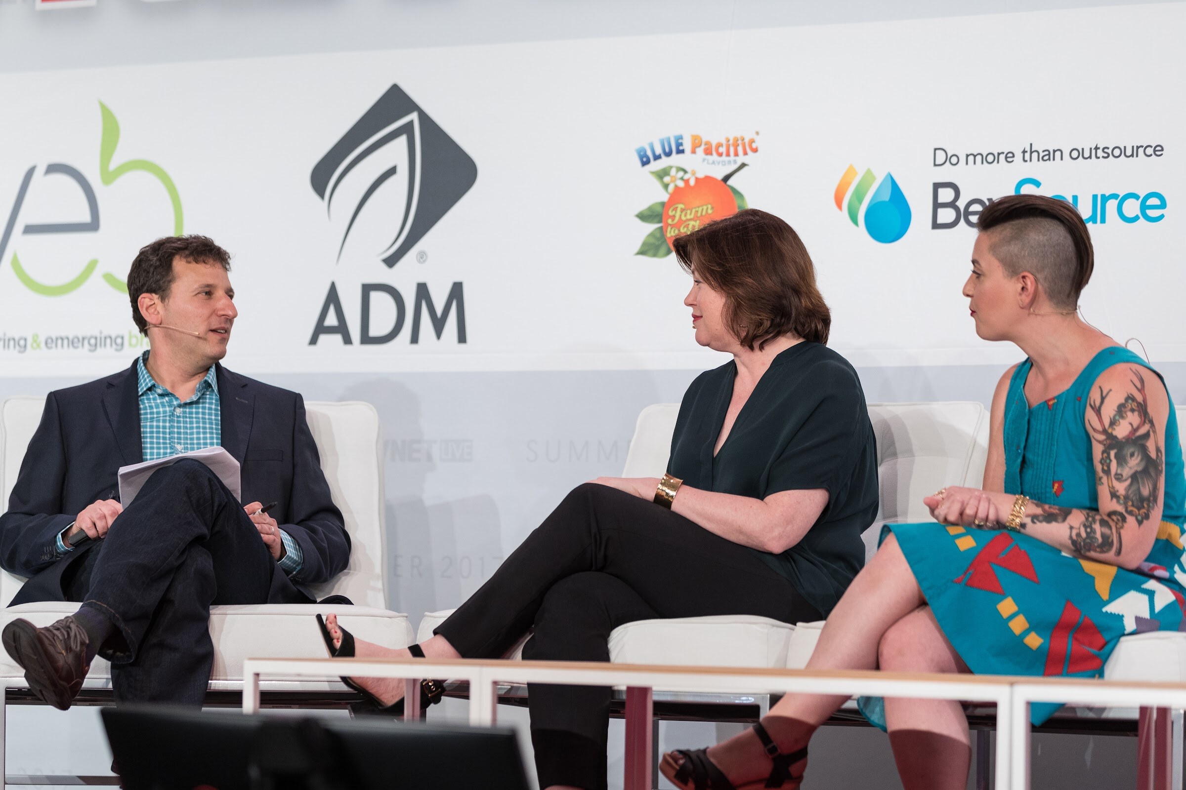 A man holding papers and two women seated on white chairs on stage, in front of VEB, ADM, Blue Pacific Flavors and BevSource logos.