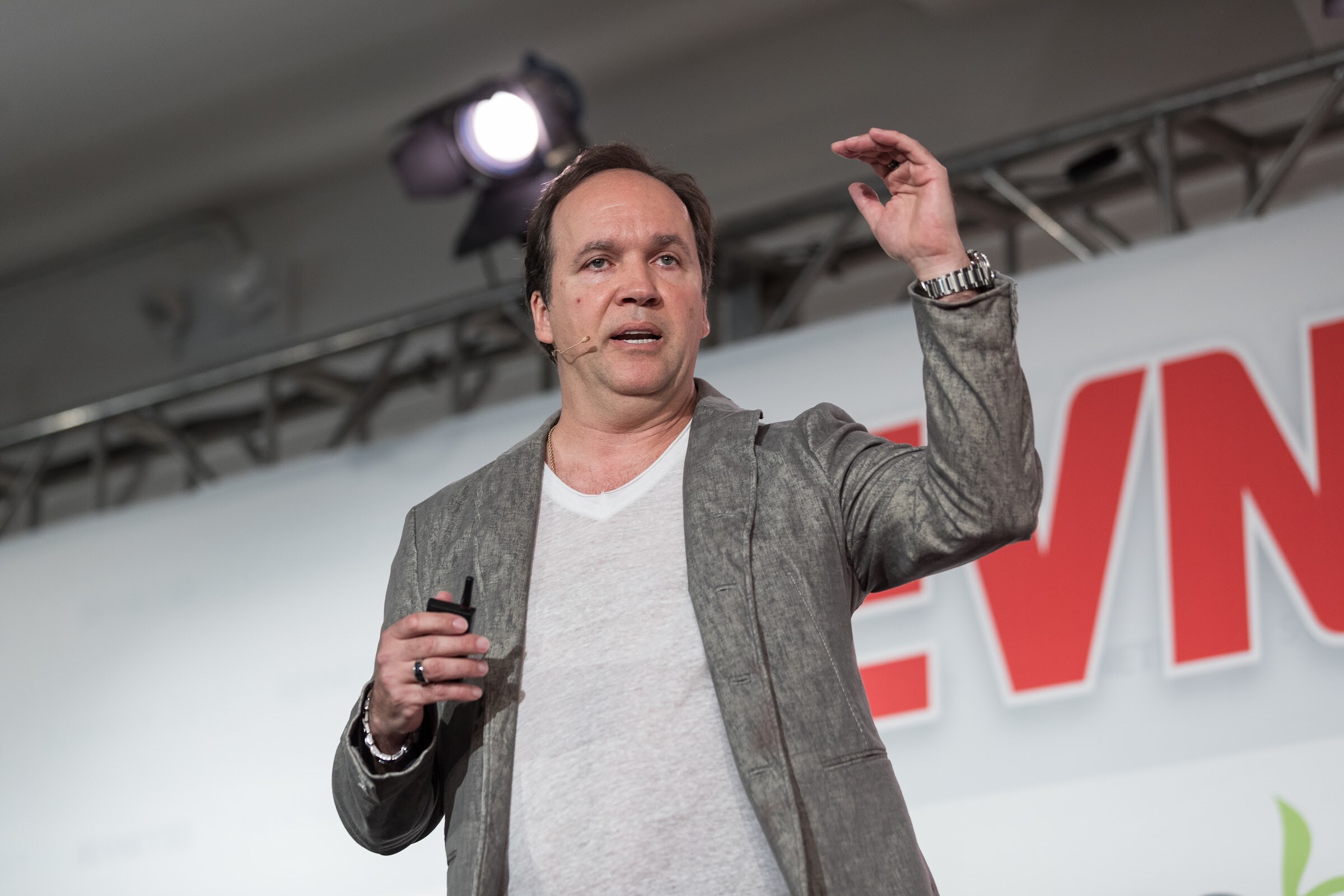 Man in a gray textured jacket and white V-neck tee with a headset microphone, one arm extended, in front of a red BEVNET LIVE sign and an ADM logo.