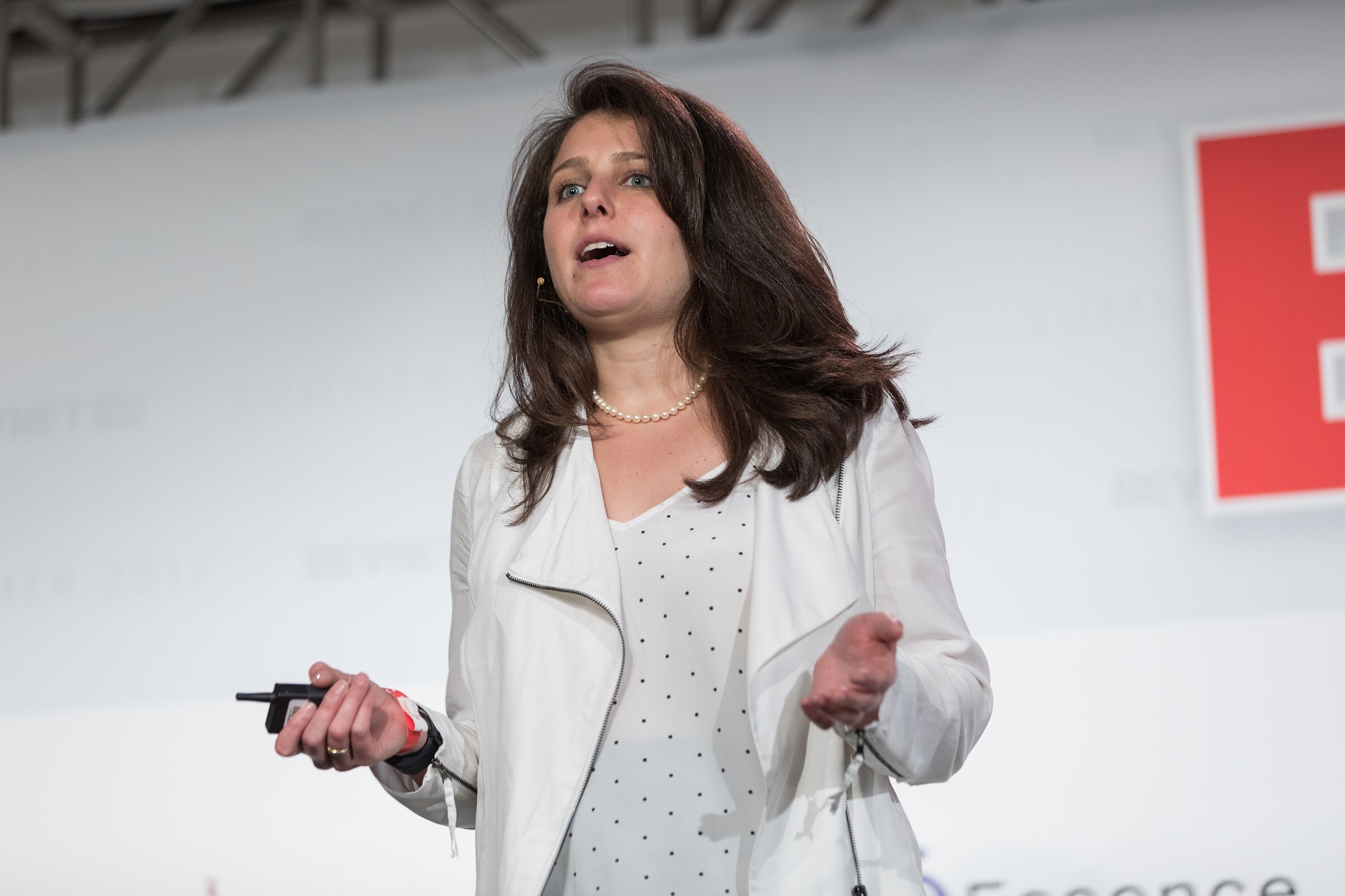 Woman with long dark hair and a pearl necklace, in a white zip jacket, speaking with a clicker in her hand against a white stage backdrop.