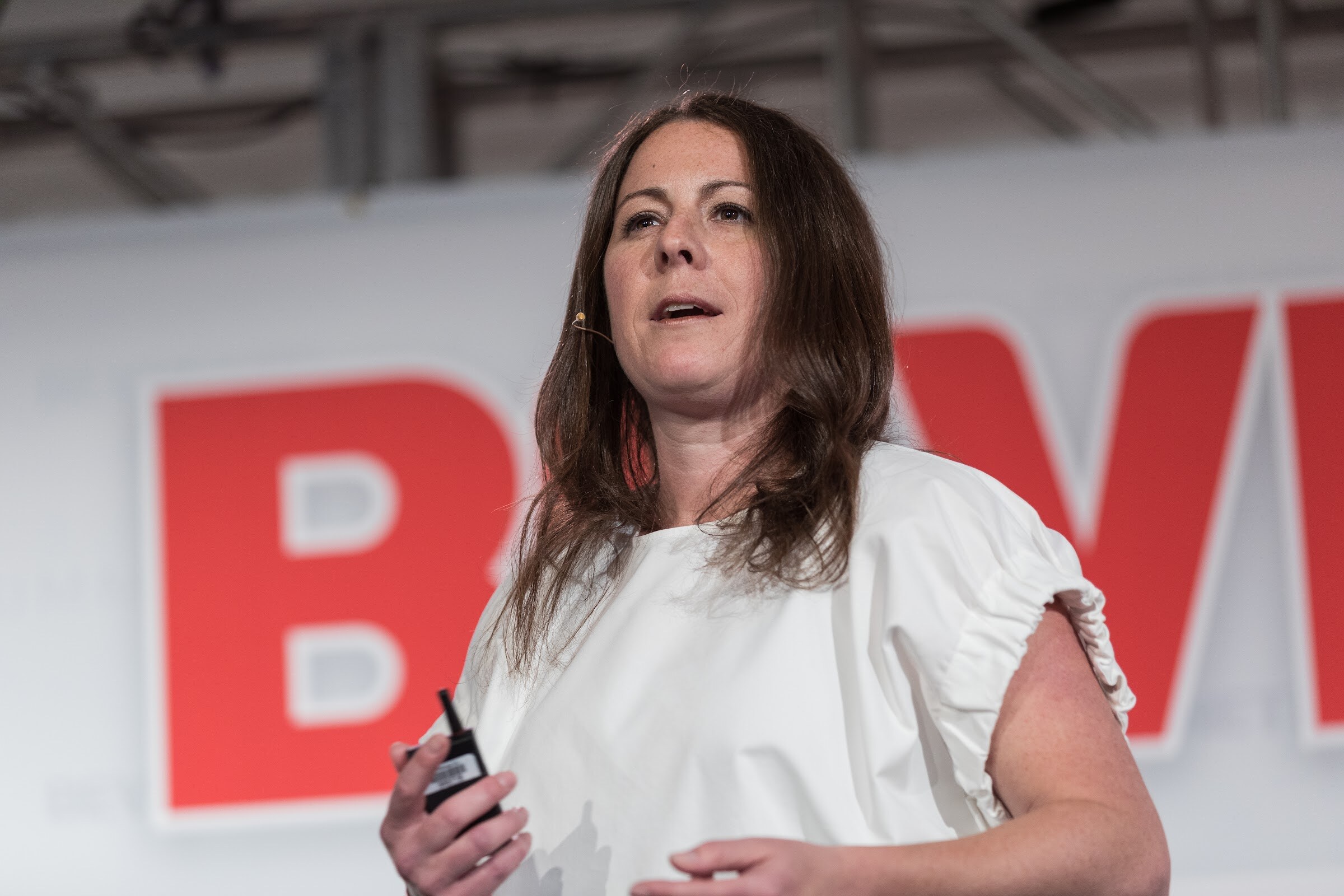 Close view of a woman with long brown hair in a white blouse speaking with a headset microphone and holding a clicker, with large red letters behind her.