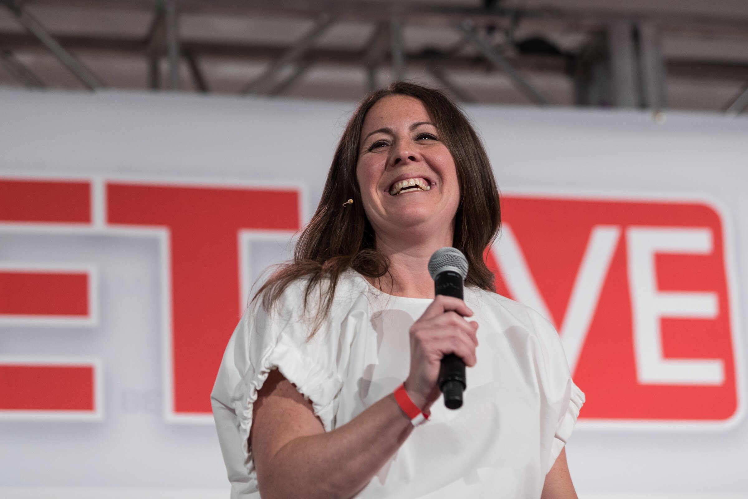 Woman in a white blouse and red wristband laughing with a black handheld microphone in front of a red BEVNET LIVE sign.