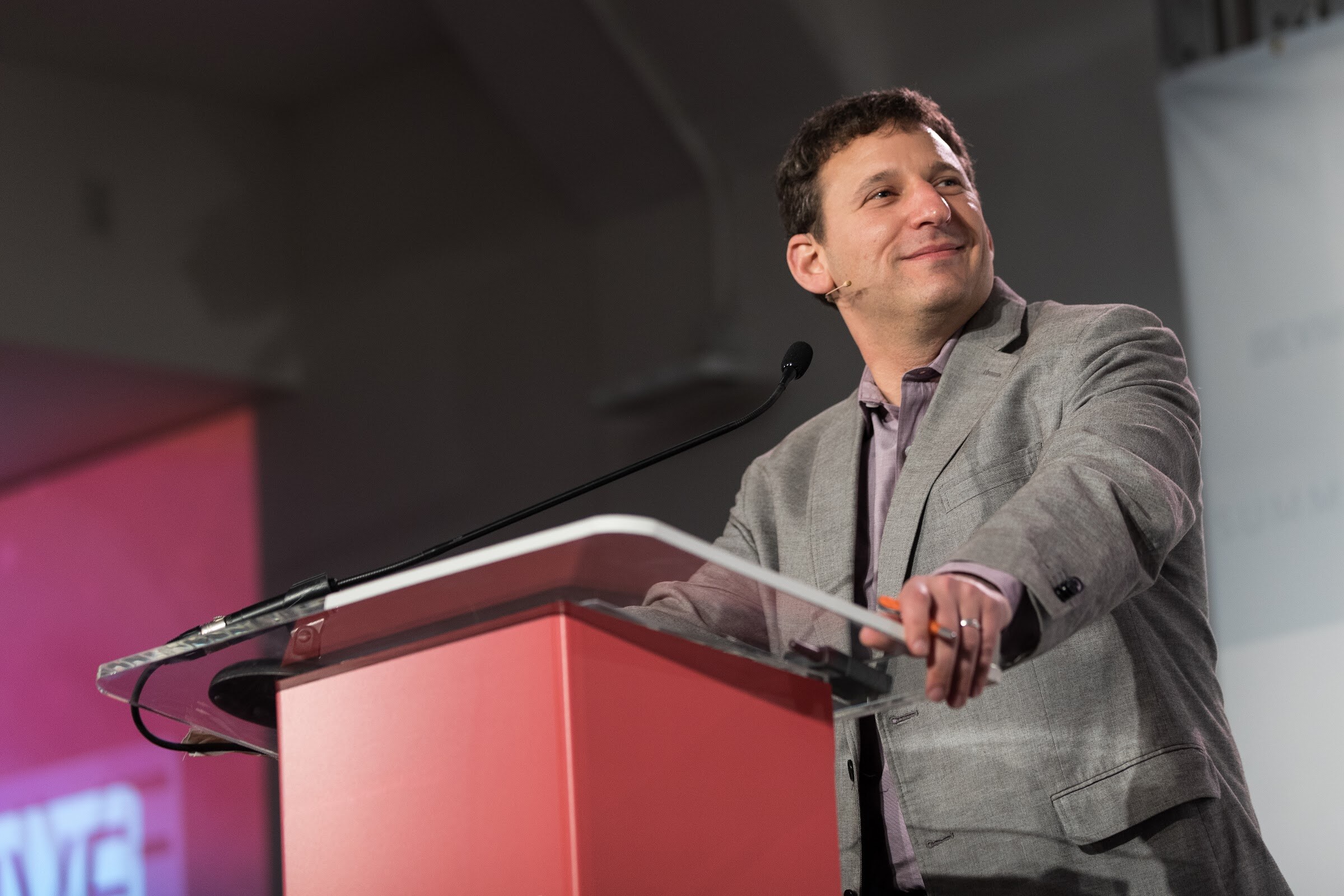 Man in a gray blazer and lavender shirt smiling at a red podium with a microphone, with a pink-lit screen behind him.