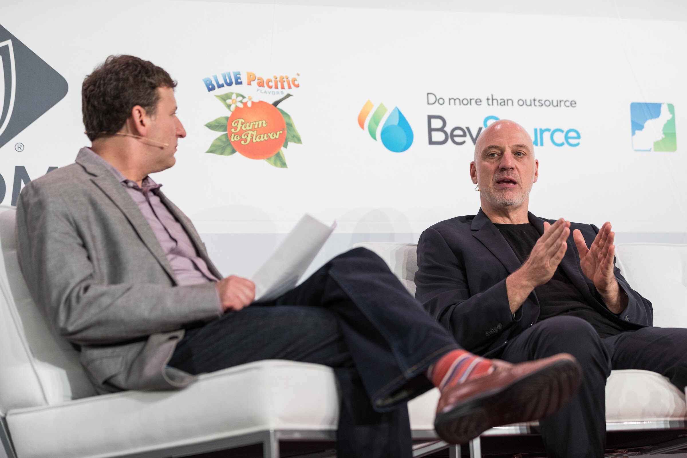 Two men on white chairs on stage; one in a gray blazer holds papers and looks at the other, who gestures with both hands, in front of Blue Pacific Flavors and BevSource logos.