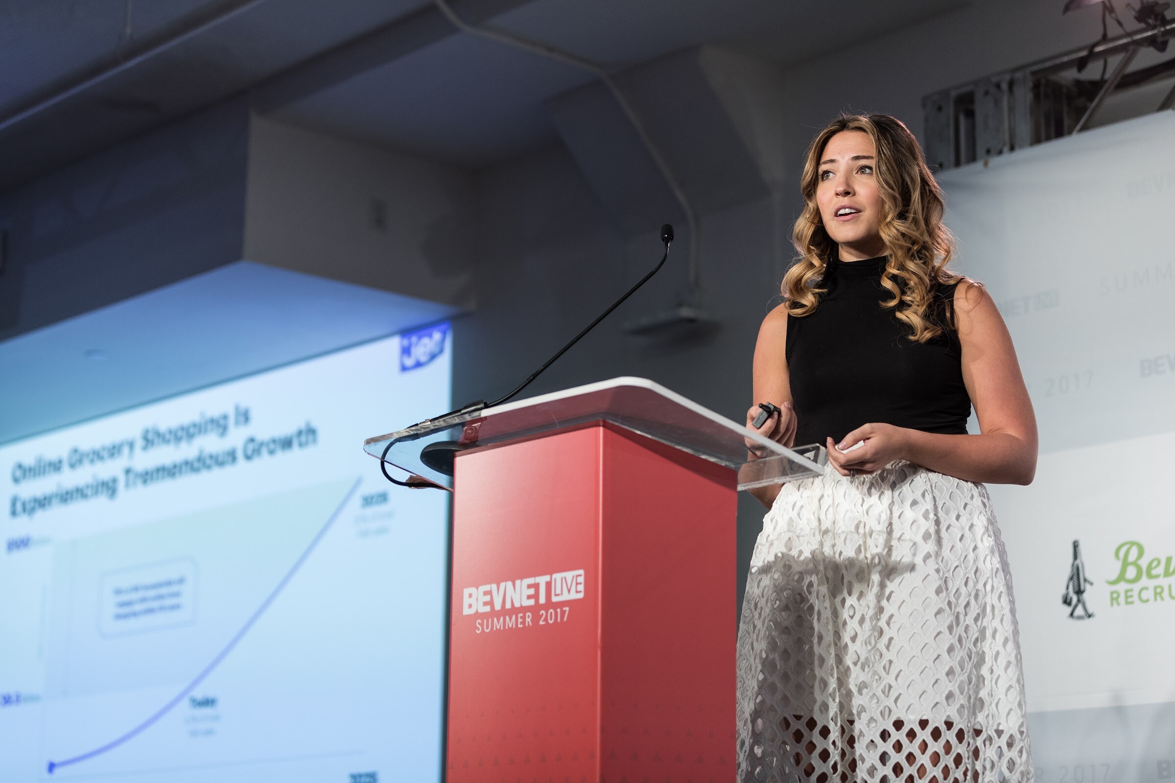 Woman with wavy hair in a black top and white cutout skirt at a BevNET Live Summer 2017 podium, with a slide headed Online Grocery Shopping Is Experiencing Tremendous Growth to her left.