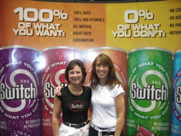 Two smiling women stand with arms around each other in front of a banner of The Switch juice cans reading "100% of what you want, 0% of what you don't."