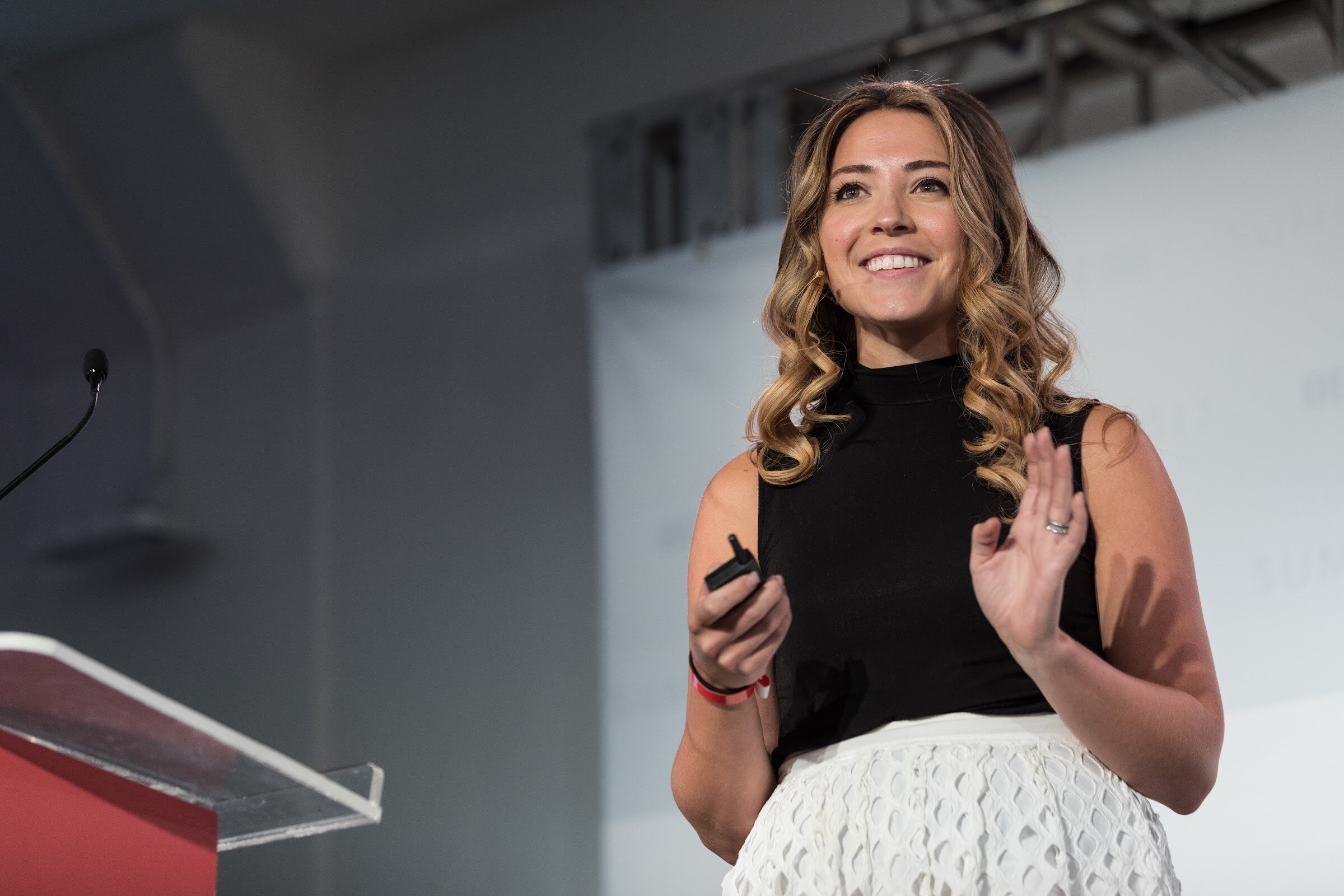 Smiling woman with wavy hair in a black sleeveless top holding a clicker and raising one hand beside a clear podium.