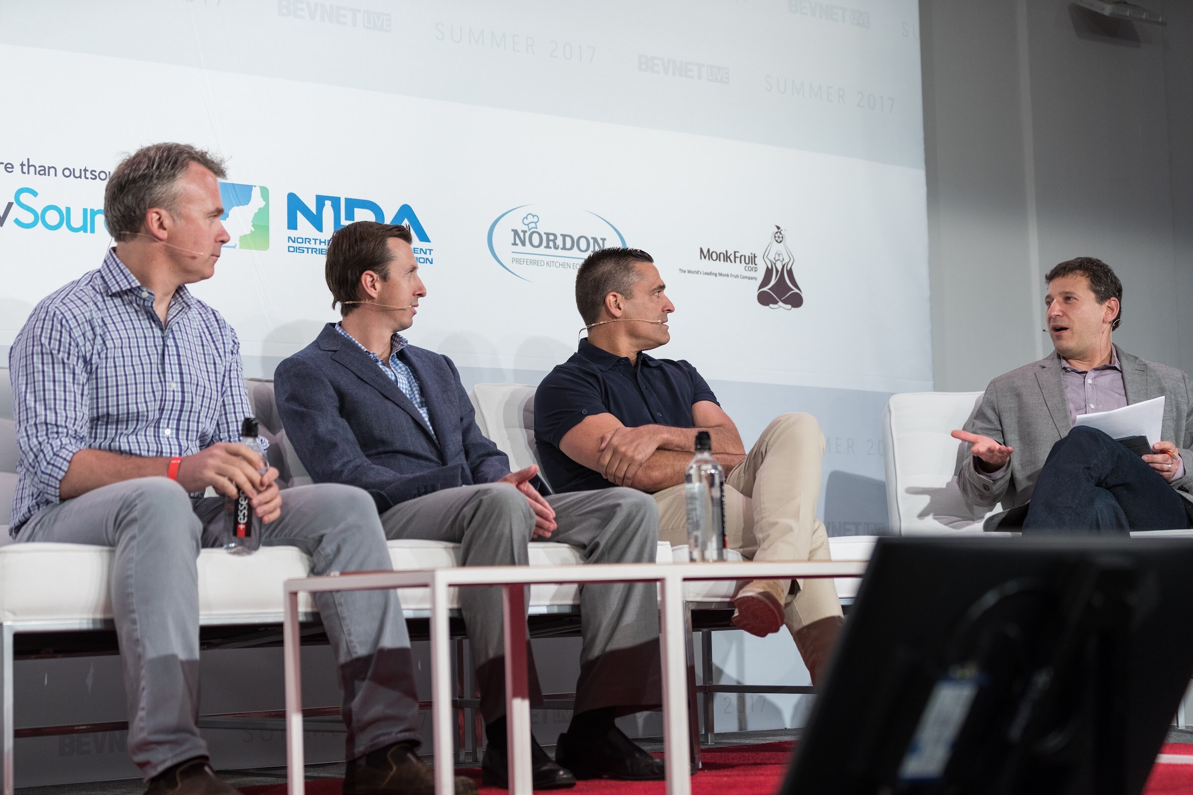 Three men seated on white chairs on stage looking right toward a fourth man holding papers and speaking, in front of NIDA, Nordon and Monk Fruit logos with a red carpet and monitor in the foreground.