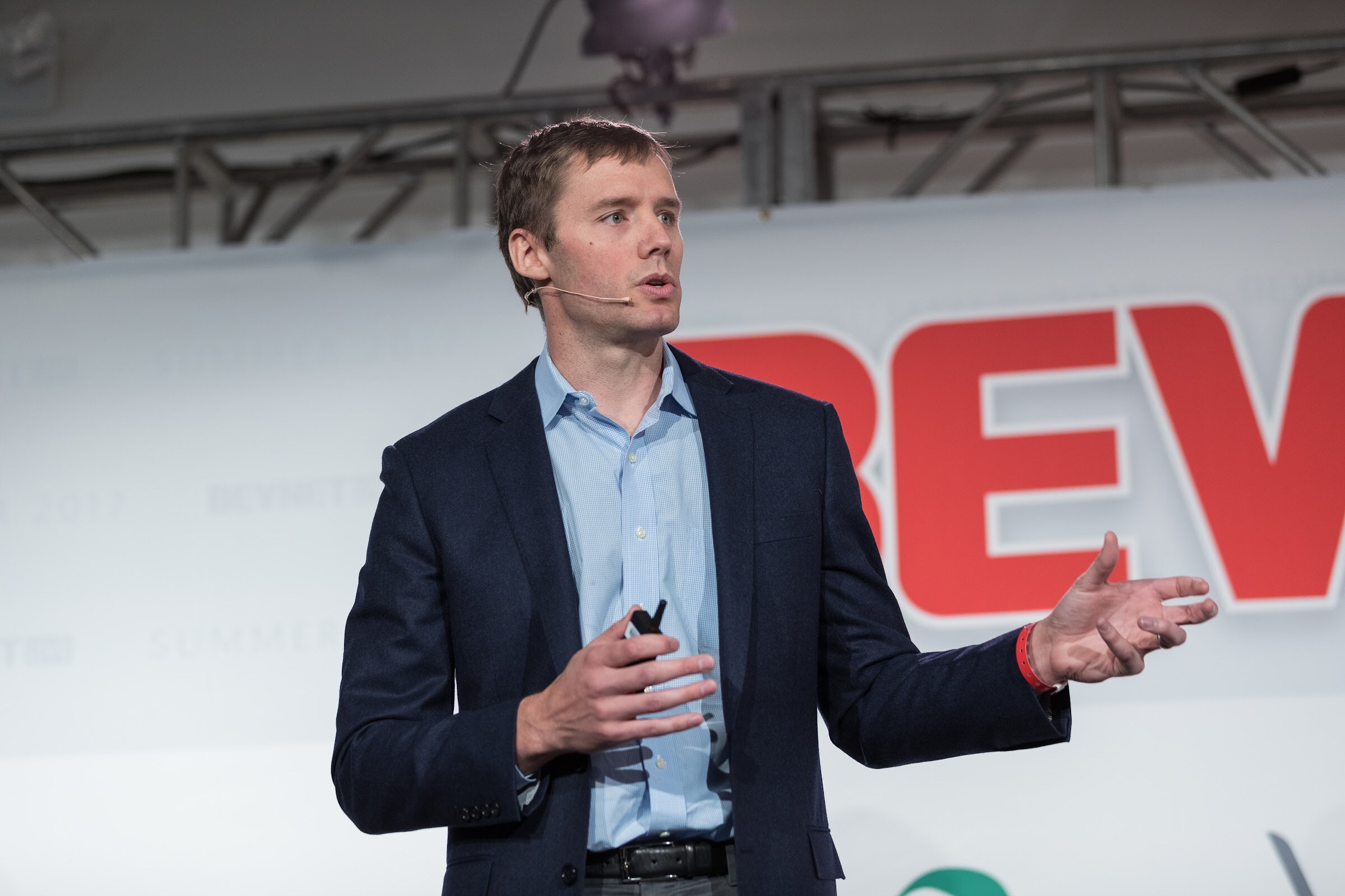 Man in a navy blazer and light blue shirt holding a clicker and gesturing with one hand in front of a red BEVNET sign.