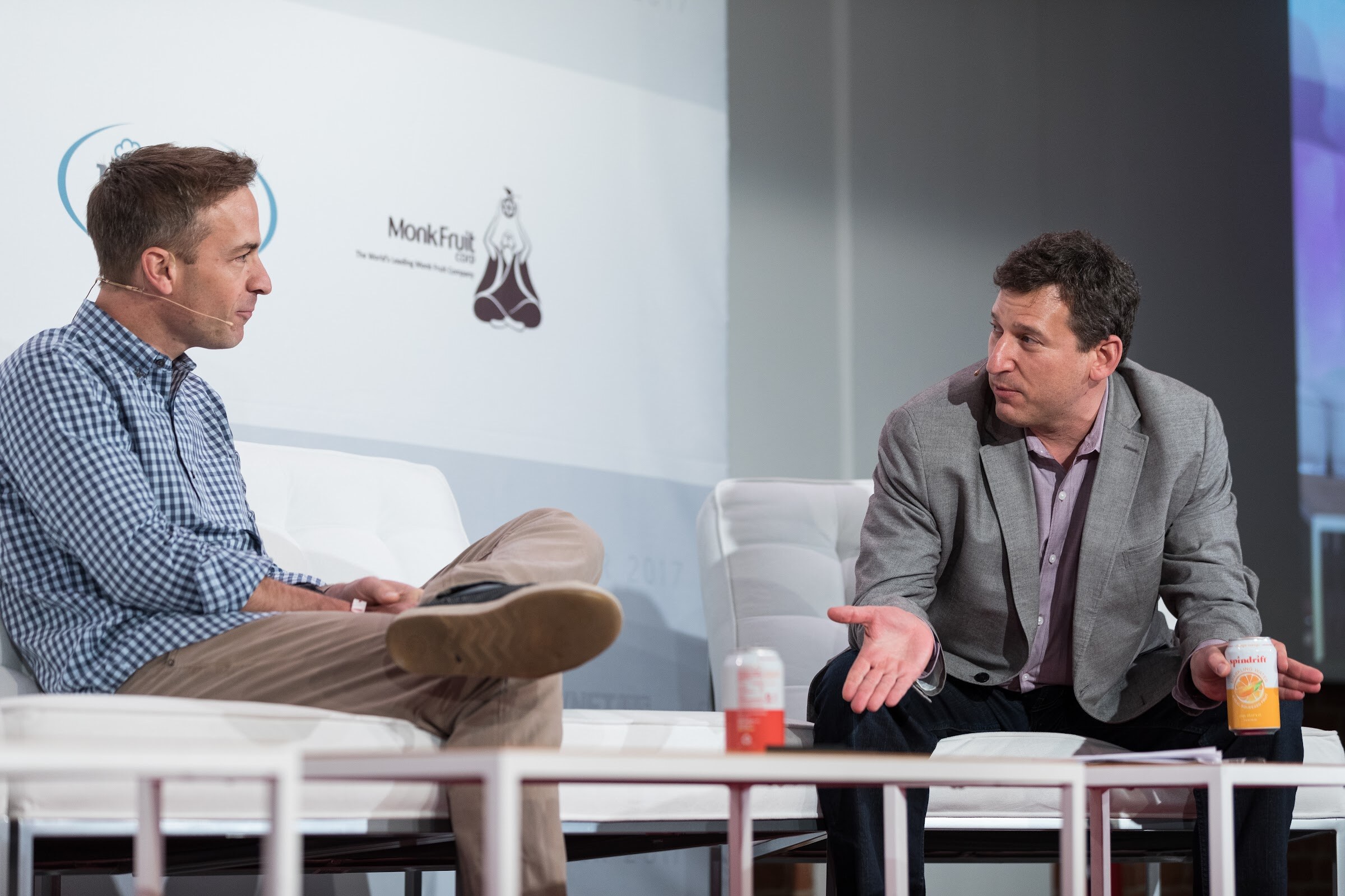 Two men on white chairs on stage; the one at right in a gray blazer holds a can of sparkling juice and gestures toward the man at left in a gingham shirt, in front of a Monk Fruit logo.