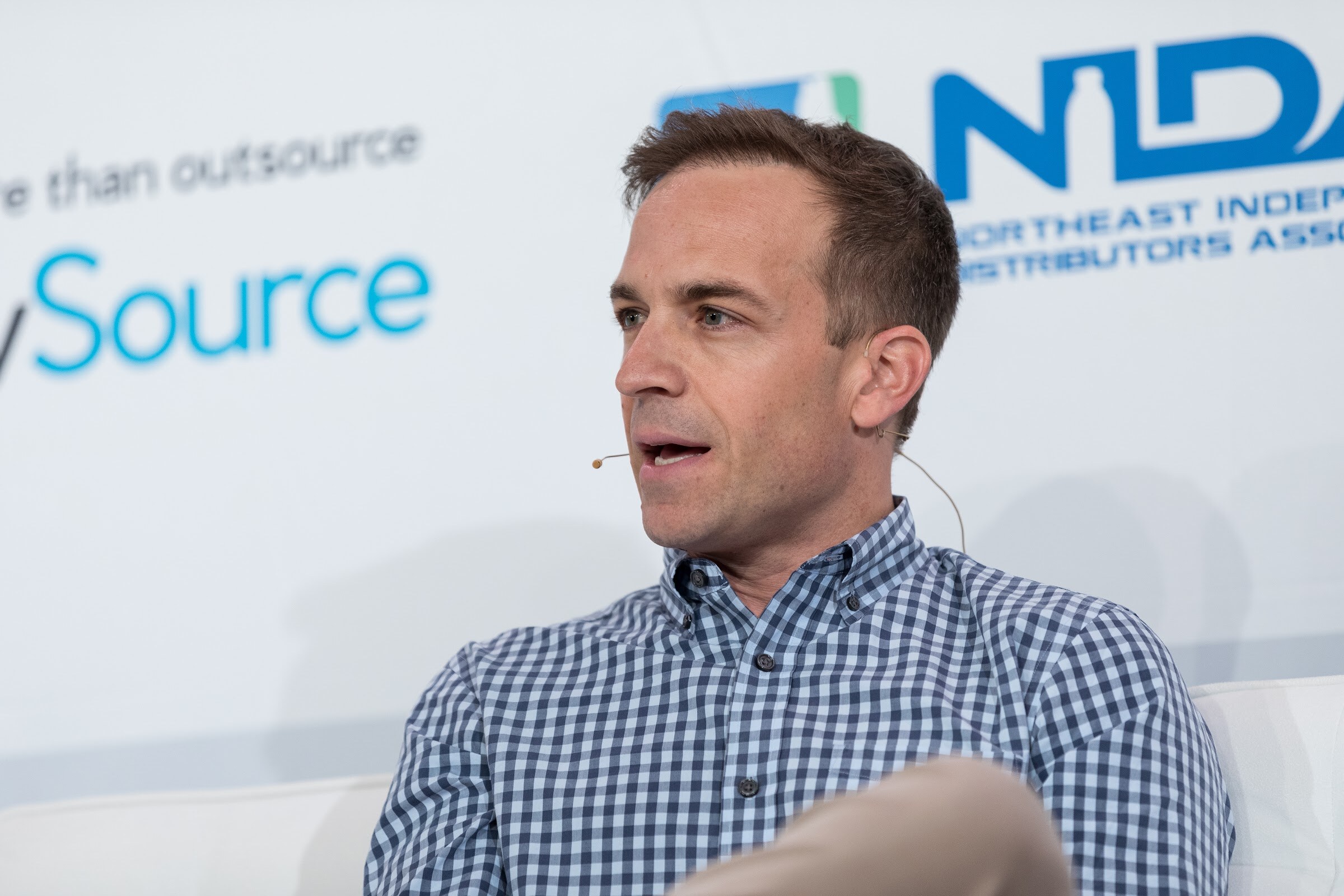 Close view of a man in a blue gingham shirt speaking with a headset microphone in front of BevSource and NIDA logos.