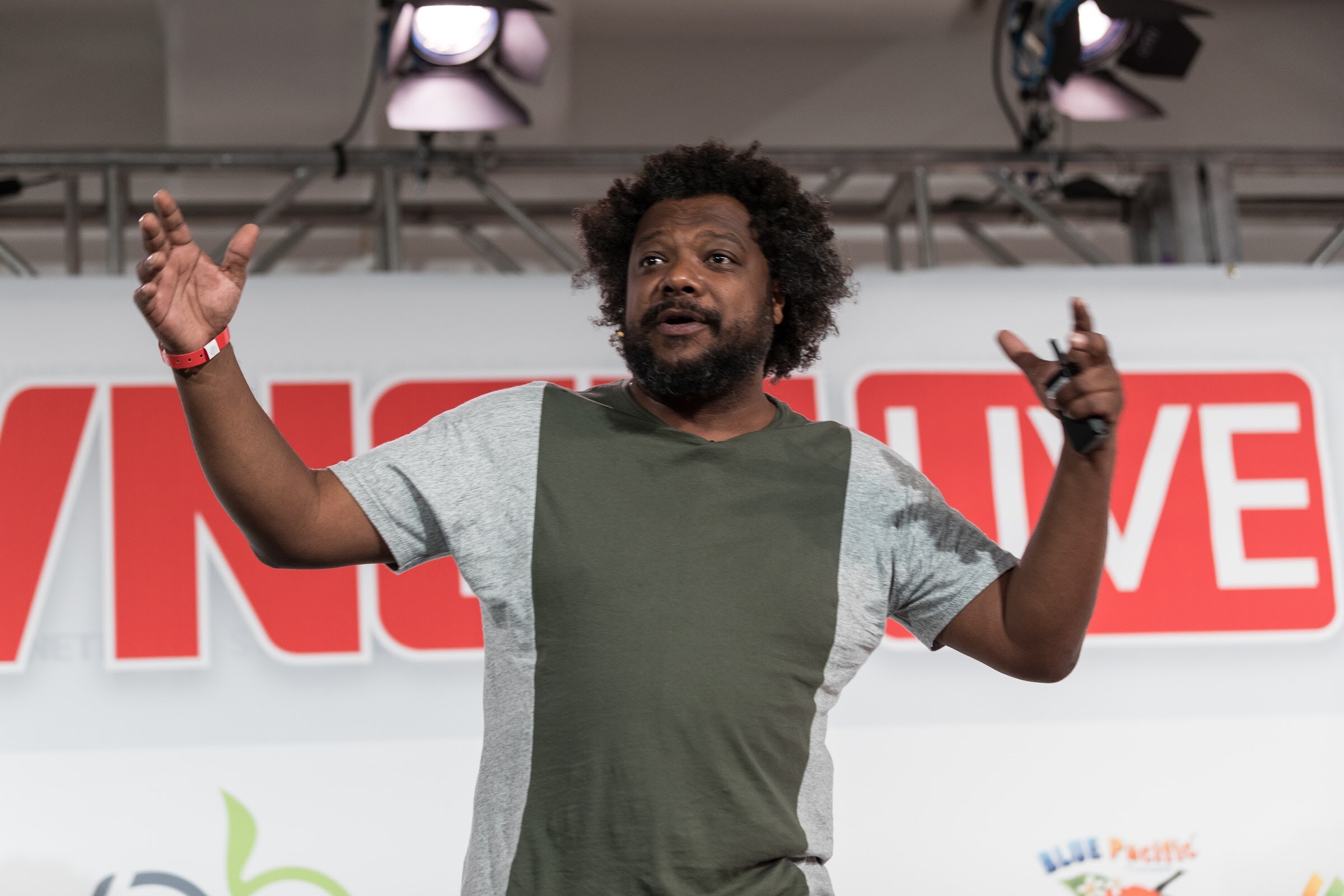 Bearded man with curly hair in a green and gray shirt, arms raised, holding a clicker in front of a red BEVNET LIVE sign and stage lights.