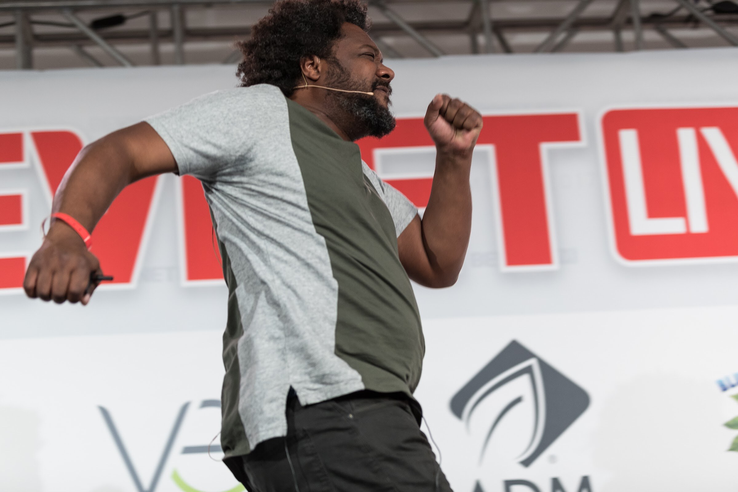 Man in a green and gray shirt leaning into a stride with one fist raised in front of a red BEVNET LIVE sign and VEB and ADM logos.