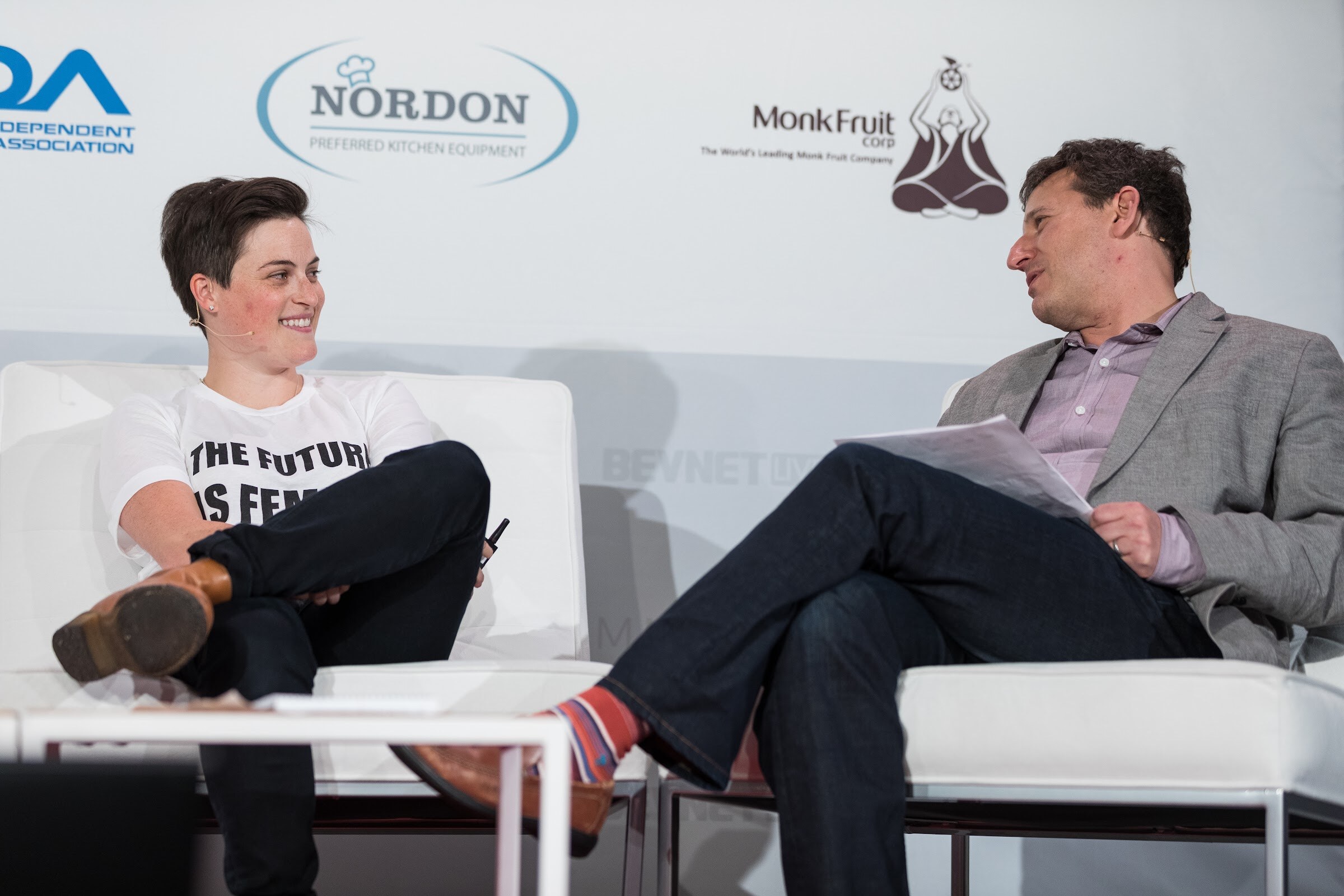 A person with short dark hair in a white T-shirt reading THE FUTURE IS FEMALE smiling at a man in a gray blazer holding papers, both seated on white chairs in front of Nordon and Monk Fruit logos.