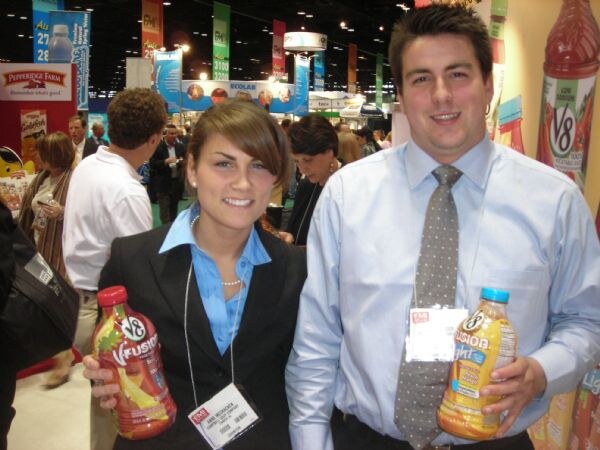 A woman in a black blazer and a man in a gray tie each hold a bottle of V8 V-Fusion juice; a Pepperidge Farm sign and a large V8 bottle graphic appear behind them.