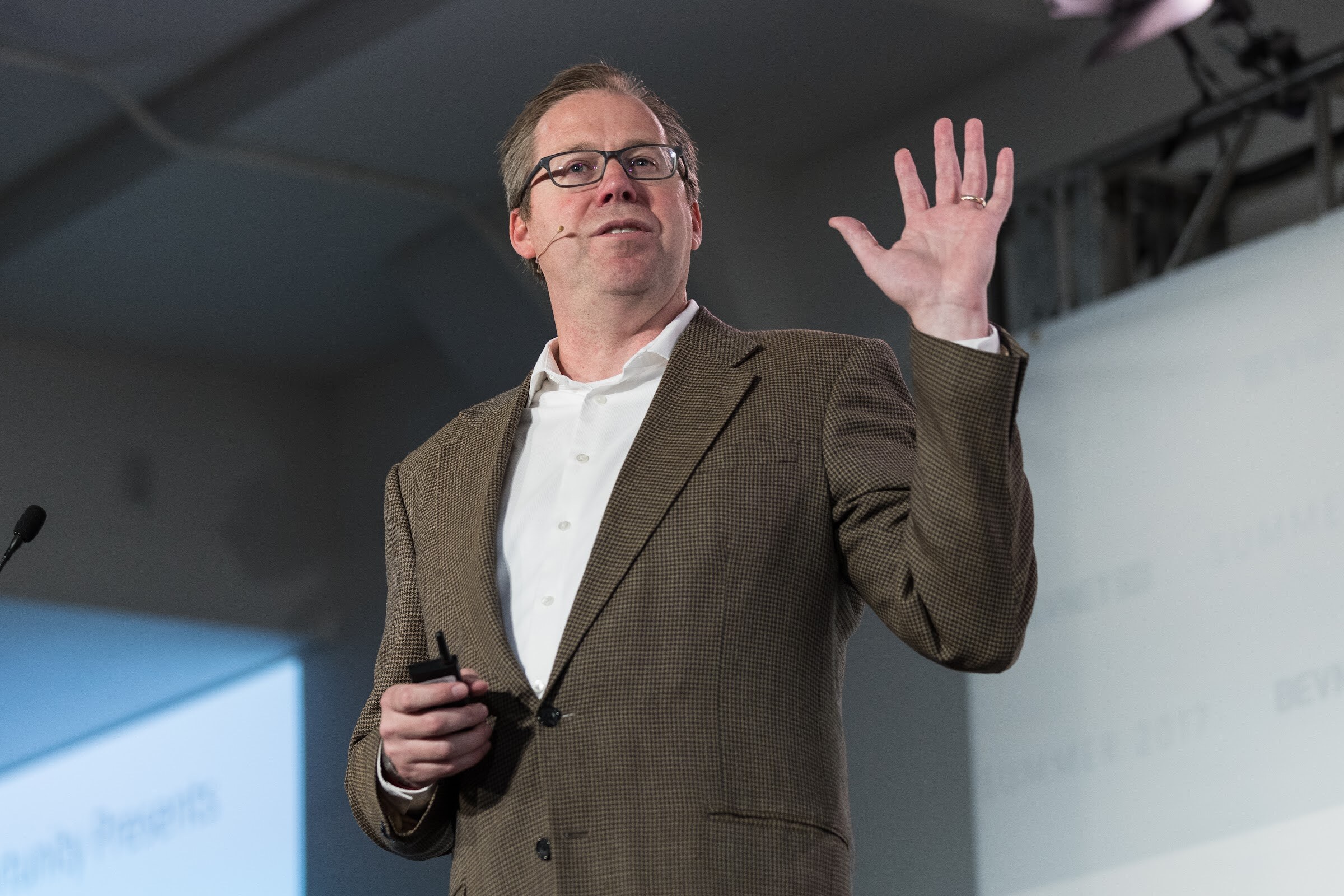Man in glasses and a brown houndstooth blazer holding a clicker and raising his right hand in front of a projection screen.