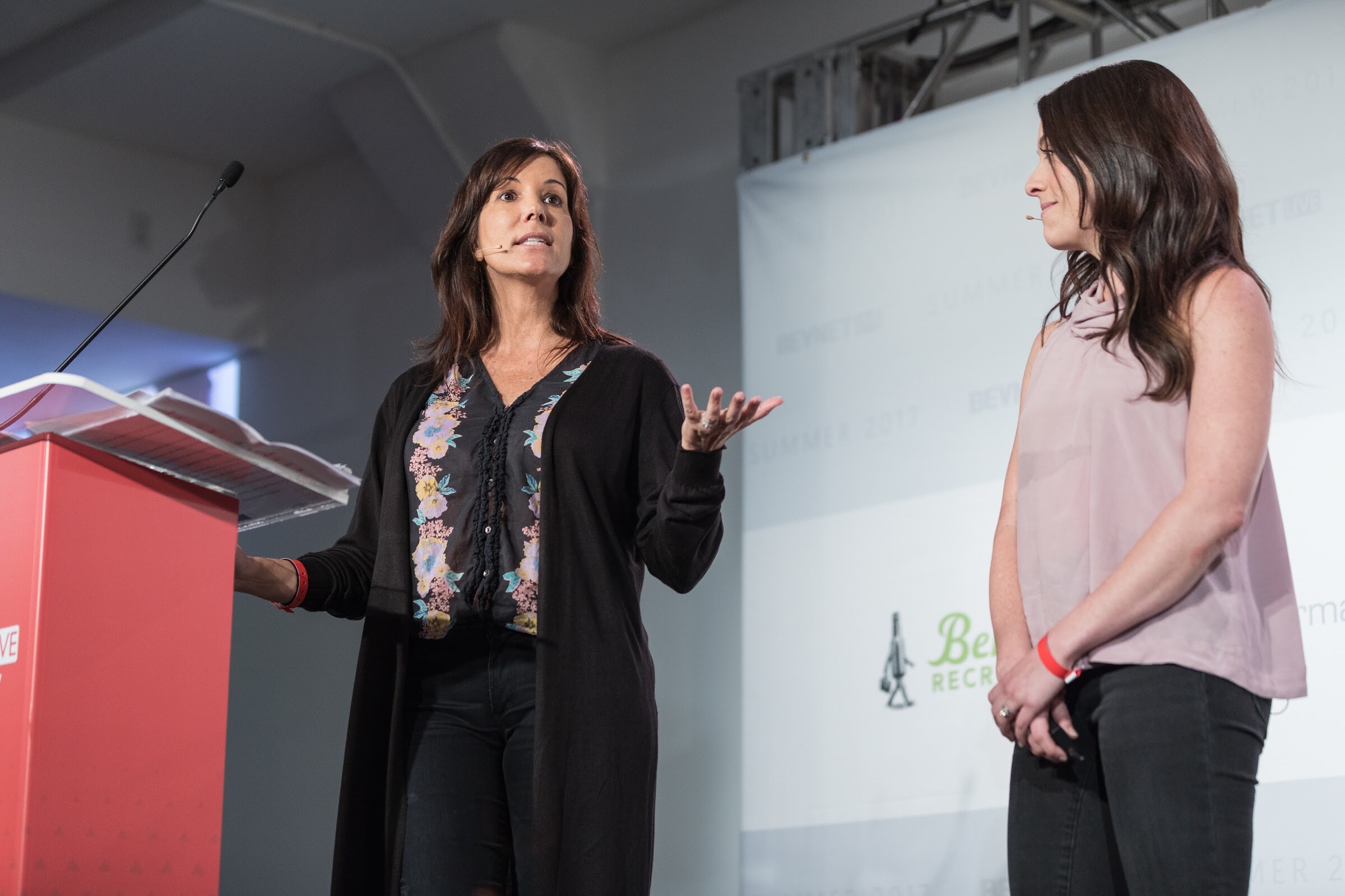 Two women on stage; one in a black cardigan with a floral top gestures and speaks beside a red podium while the other, in a pink sleeveless top, listens.