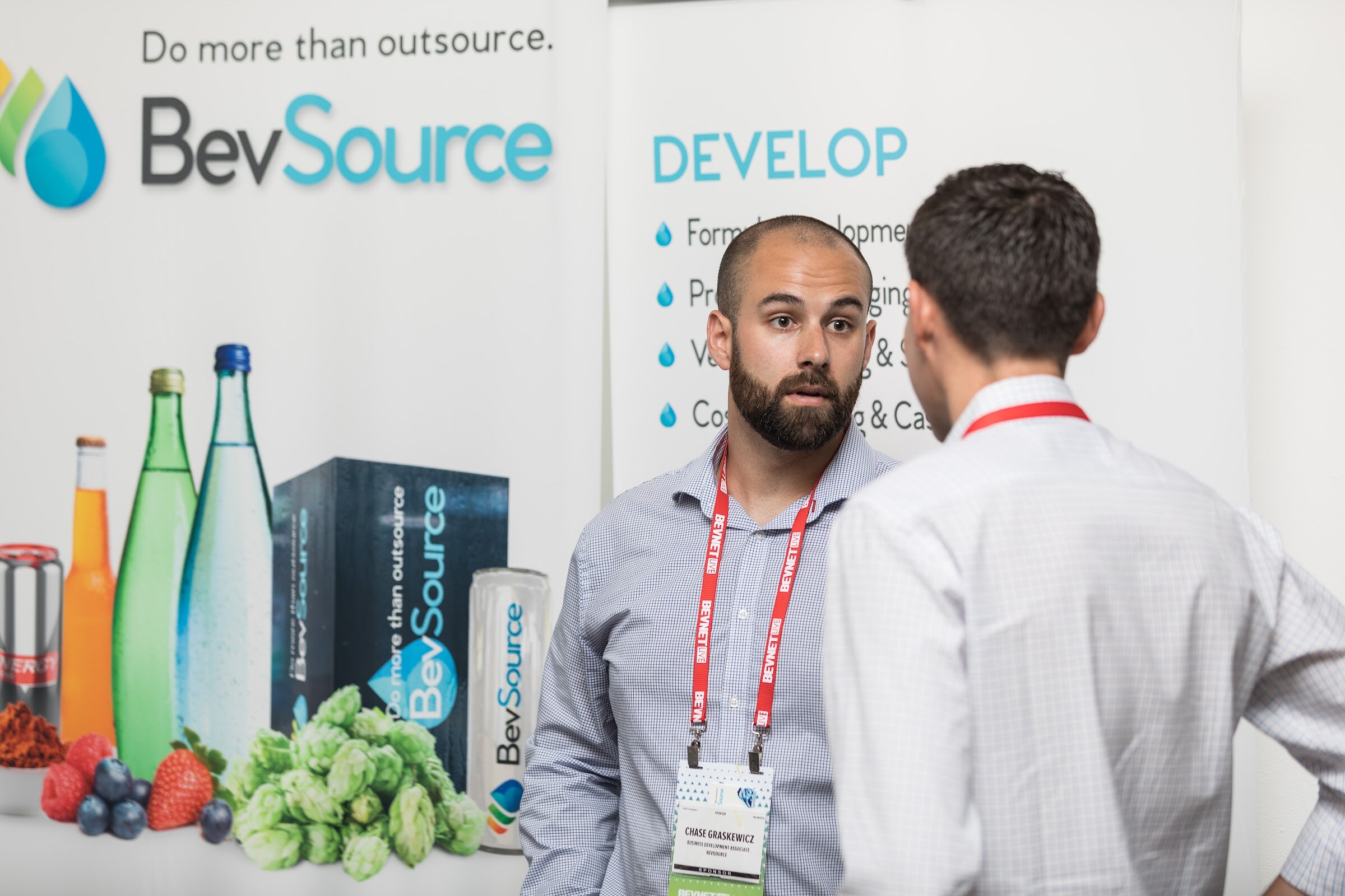 Bearded man in a gray gingham shirt and red lanyard talking with an attendee seen from behind in front of a BevSource Do more than outsource banner.