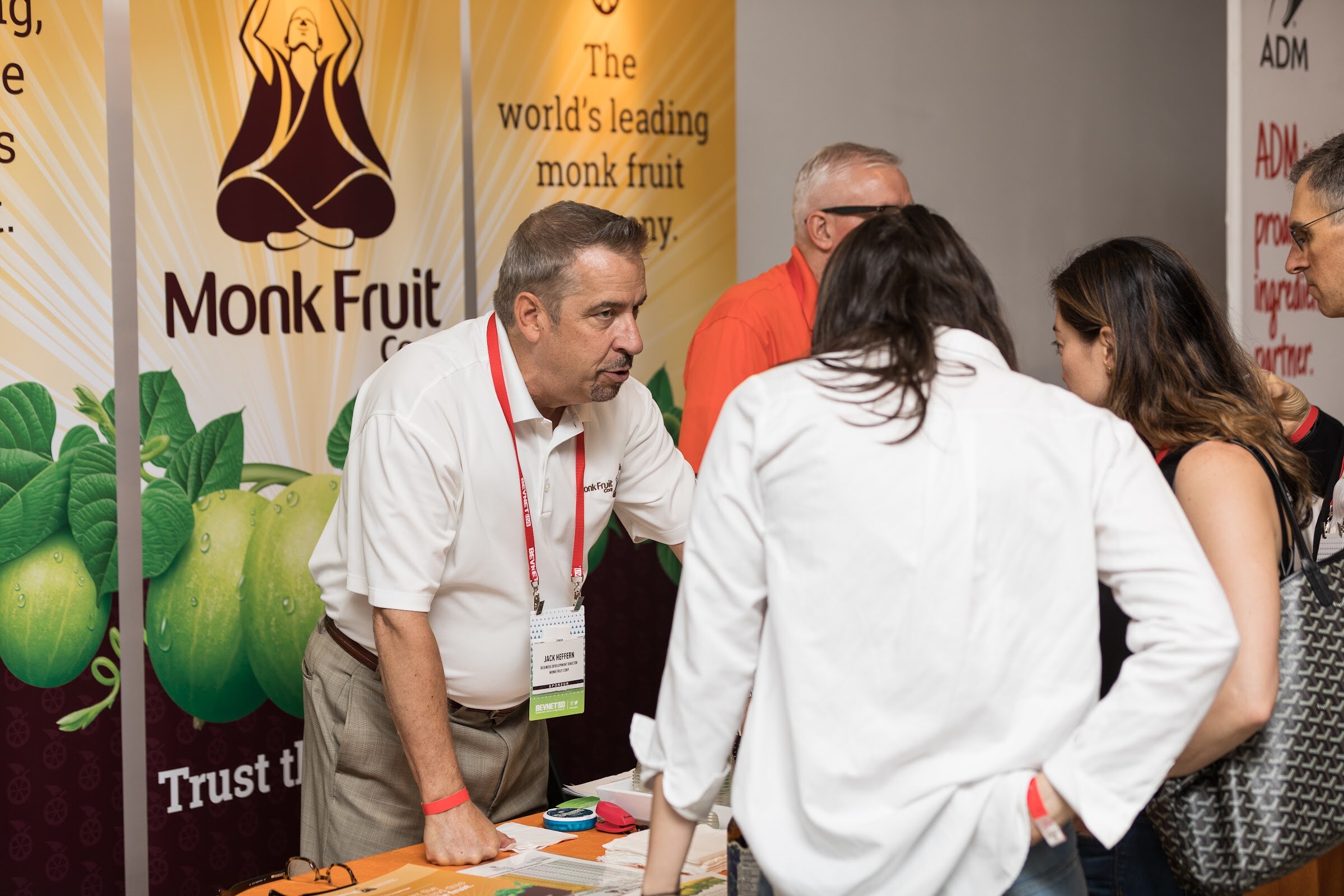 Man in a white polo and red lanyard leaning over a table to talk to attendees in front of a Monk Fruit Corp banner showing monk fruit.