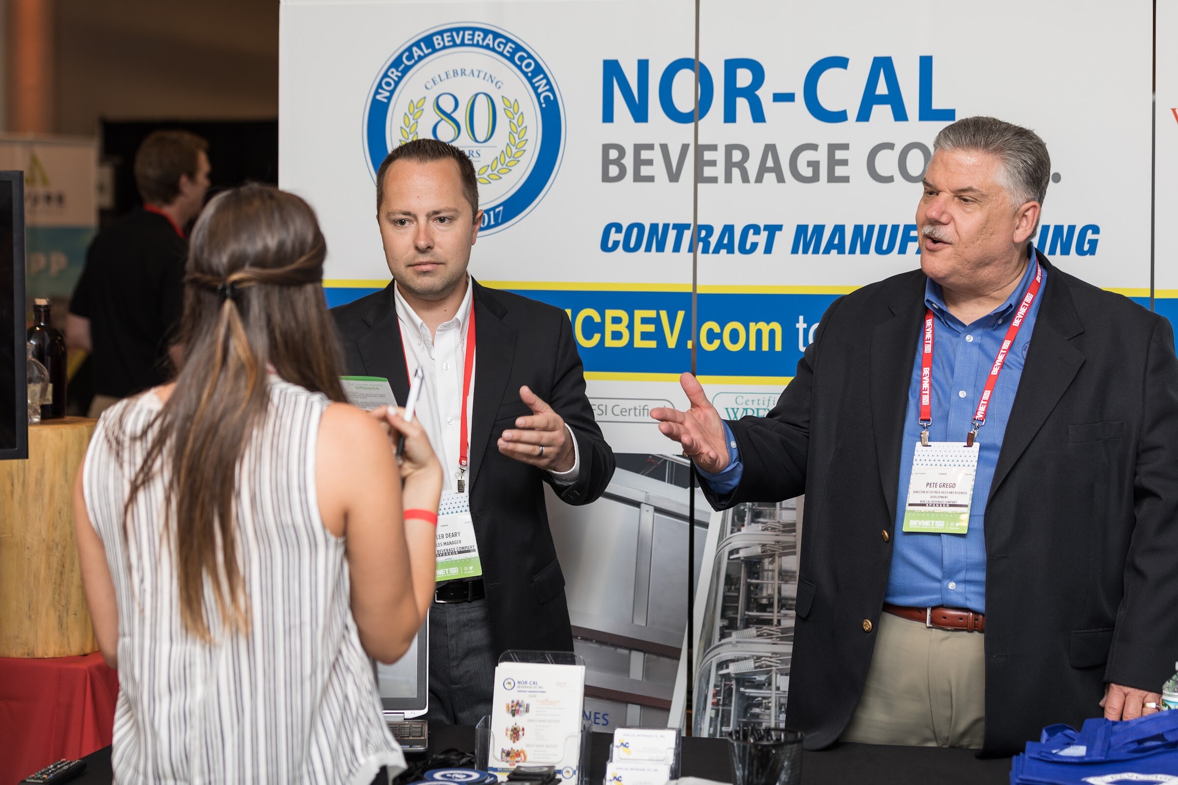 Two men in dark blazers gesturing and talking with a woman in a striped top in front of a Nor-Cal Beverage Co. contract manufacturing banner.