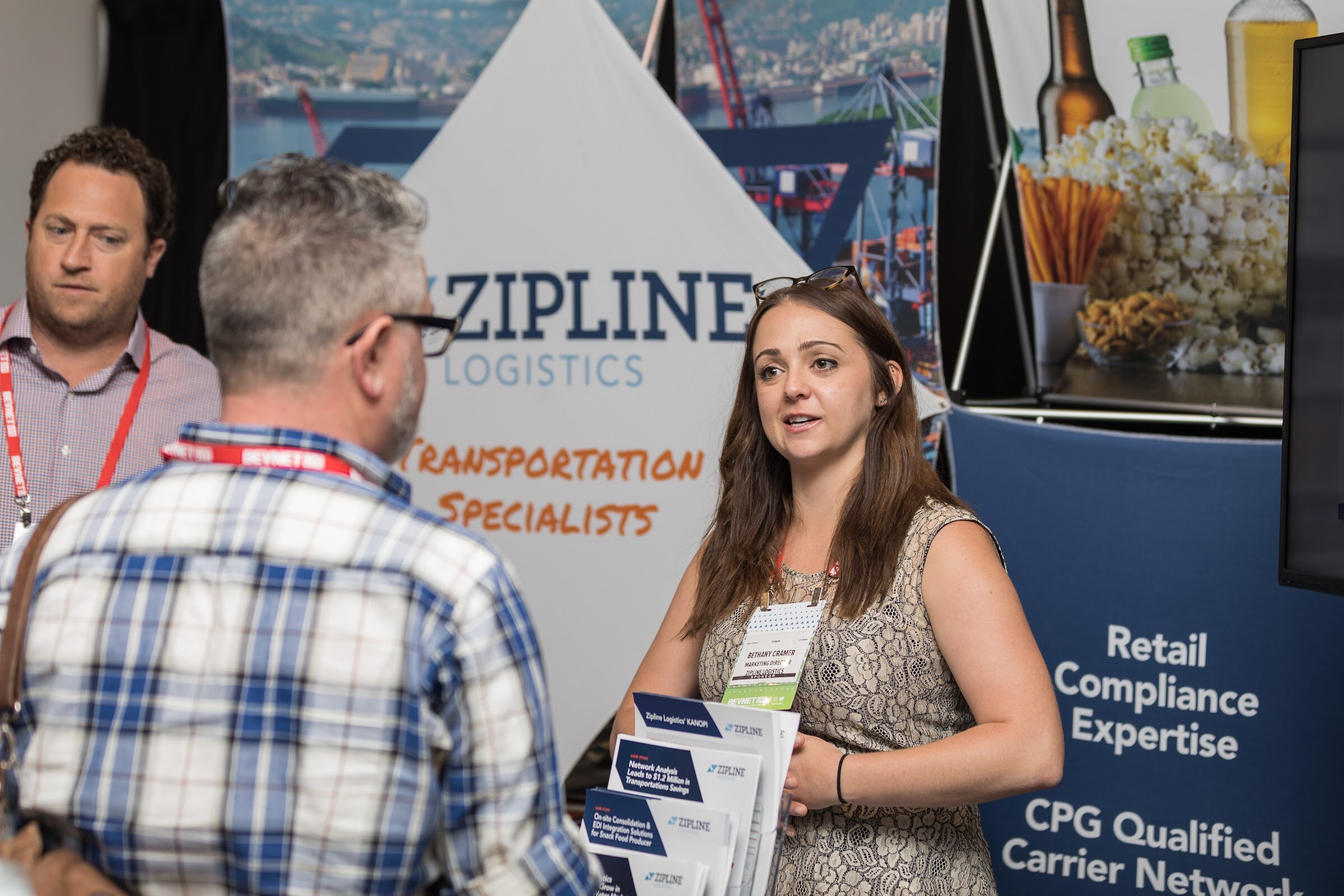 Woman with long brown hair in a lace top holding brochures and speaking with a man in a blue plaid shirt in front of a Zipline Logistics banner.