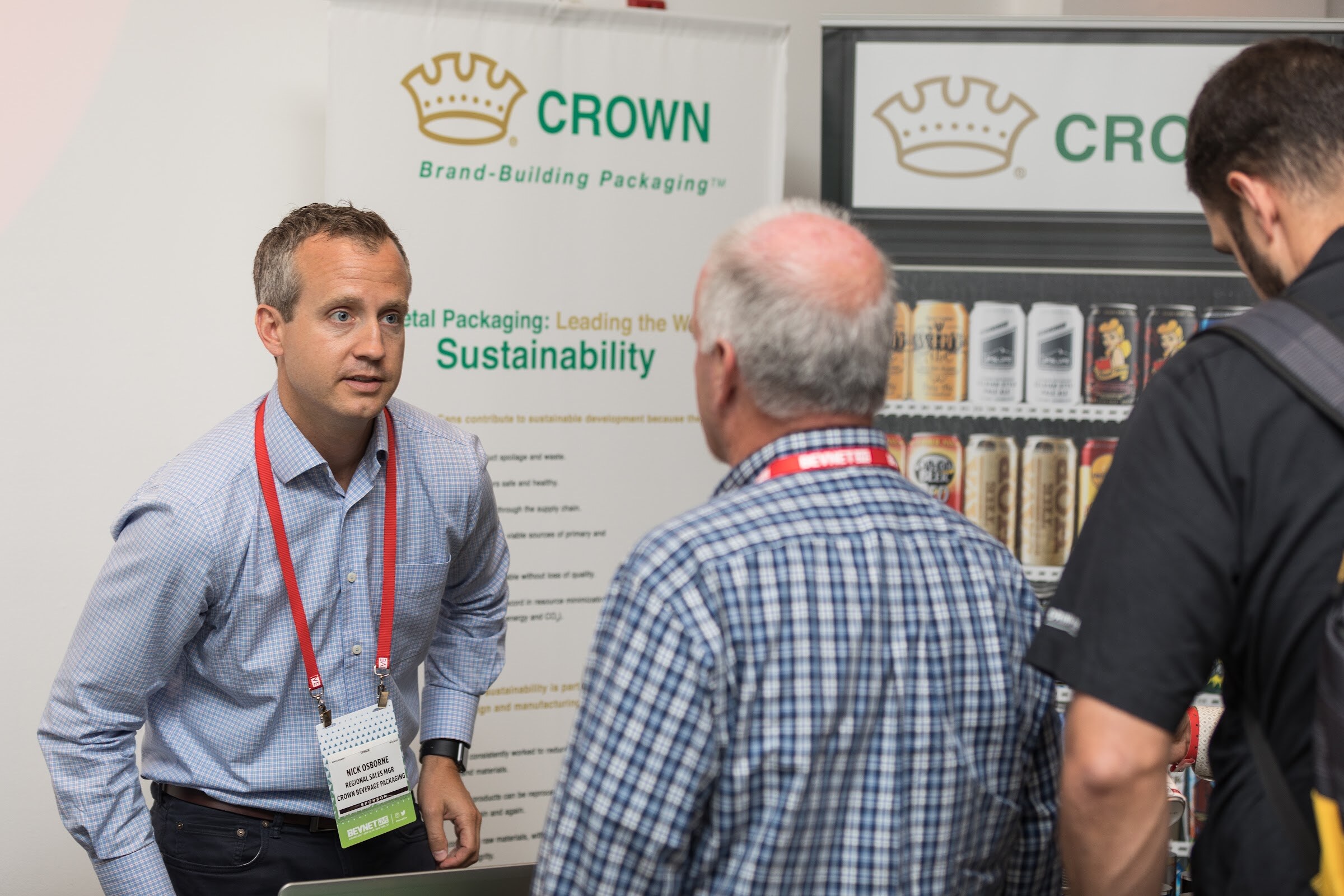 Man in a light blue shirt and red lanyard talking with two attendees in front of Crown Brand-Building Packaging banners showing cans.