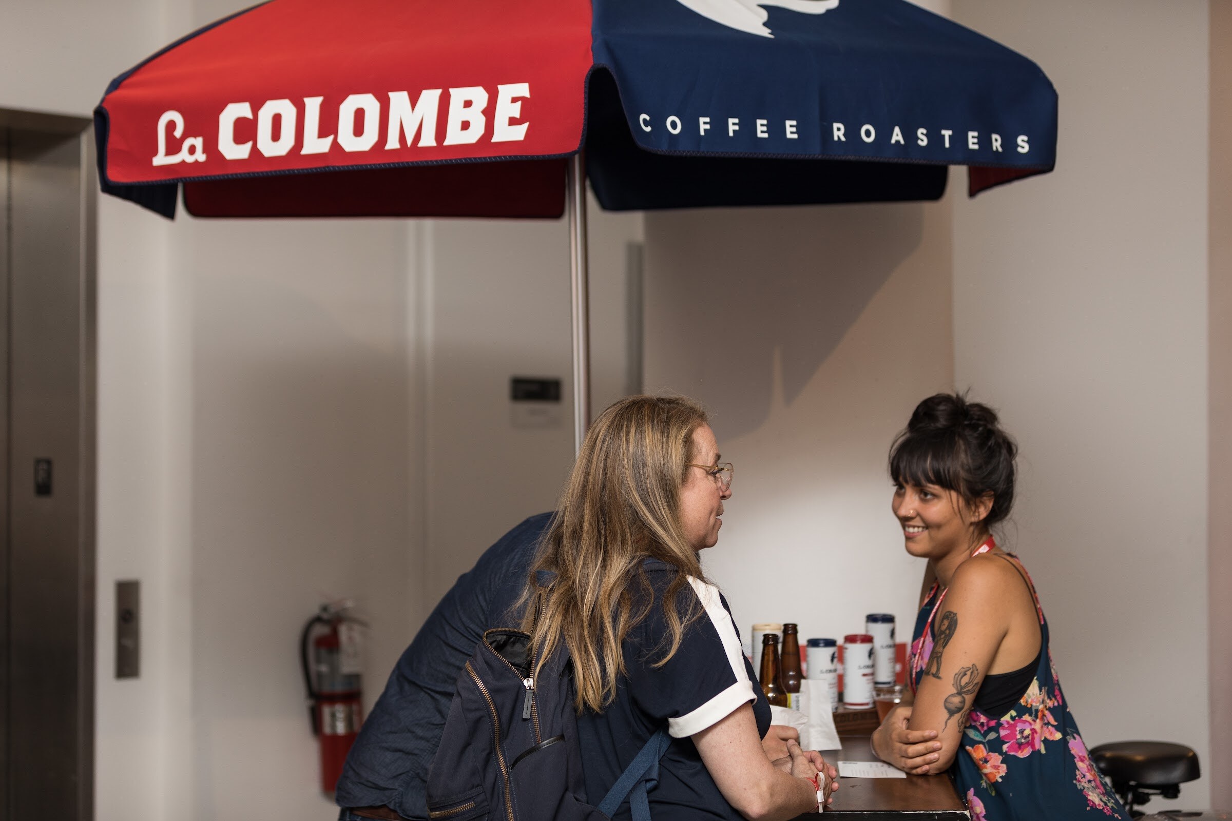 Smiling woman with a tattooed arm behind a counter talking with a woman with a backpack, under a red and navy umbrella reading La Colombe Coffee Roasters.