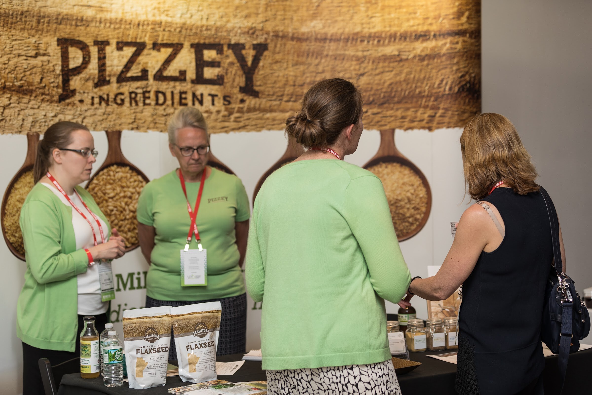 Three women in green tops and a woman with a navy bag at a booth in front of a wooden Pizzey Ingredients banner, with bags of flaxseed on the table.