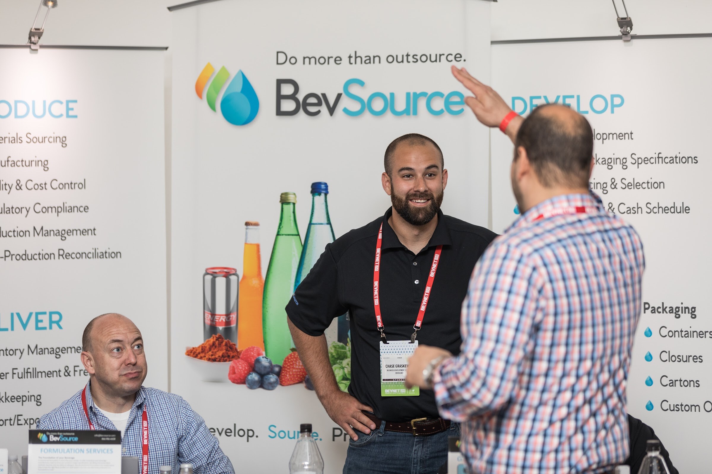 Smiling bearded man in a black polo and lanyard in front of a BevSource banner while a visitor in a plaid shirt raises a hand toward it and a seated man looks on.