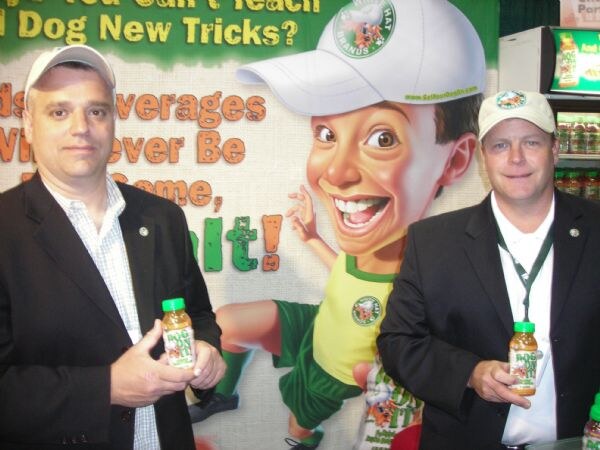 Two men in dark jackets and caps each hold a small bottle of juice in front of a White Hat Brands banner showing a cartoon boy in a white cap.