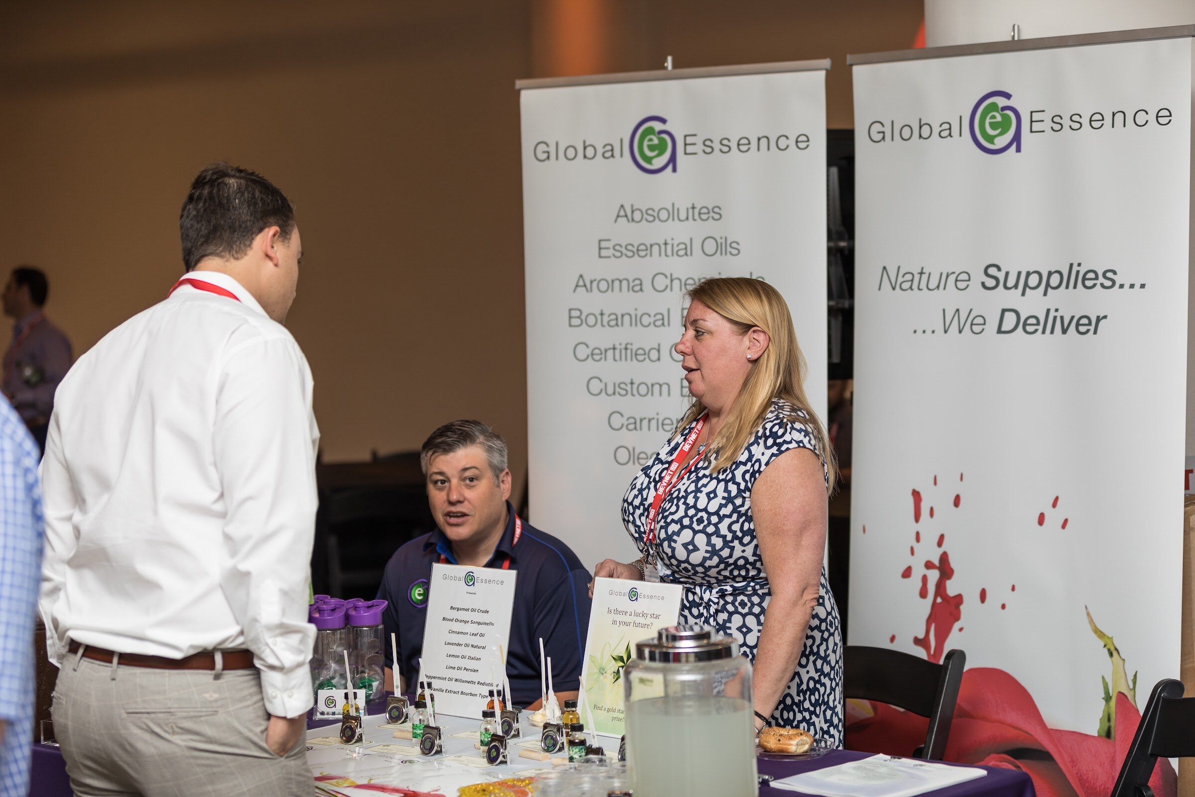 A man in a white shirt talking to a seated man in a navy polo and a standing woman in a blue patterned dress at a table with Global Essence banners.