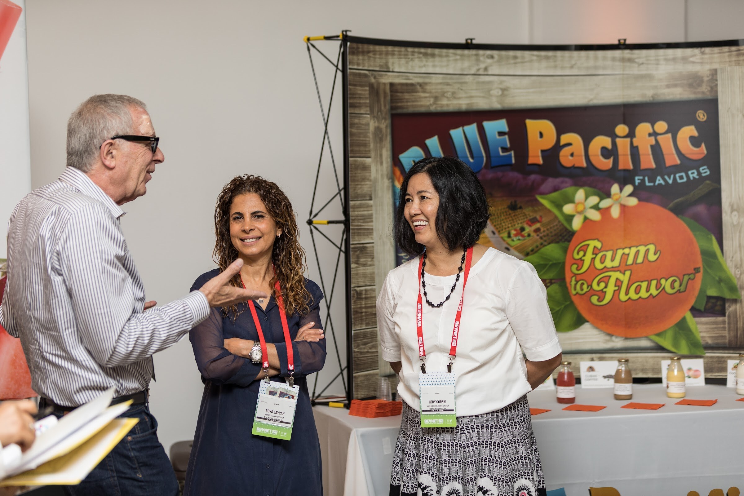 A gray-haired man in glasses talking with two smiling women in lanyards in front of a Blue Pacific Flavors Farm to Flavor backdrop with juice sample bottles on the table.