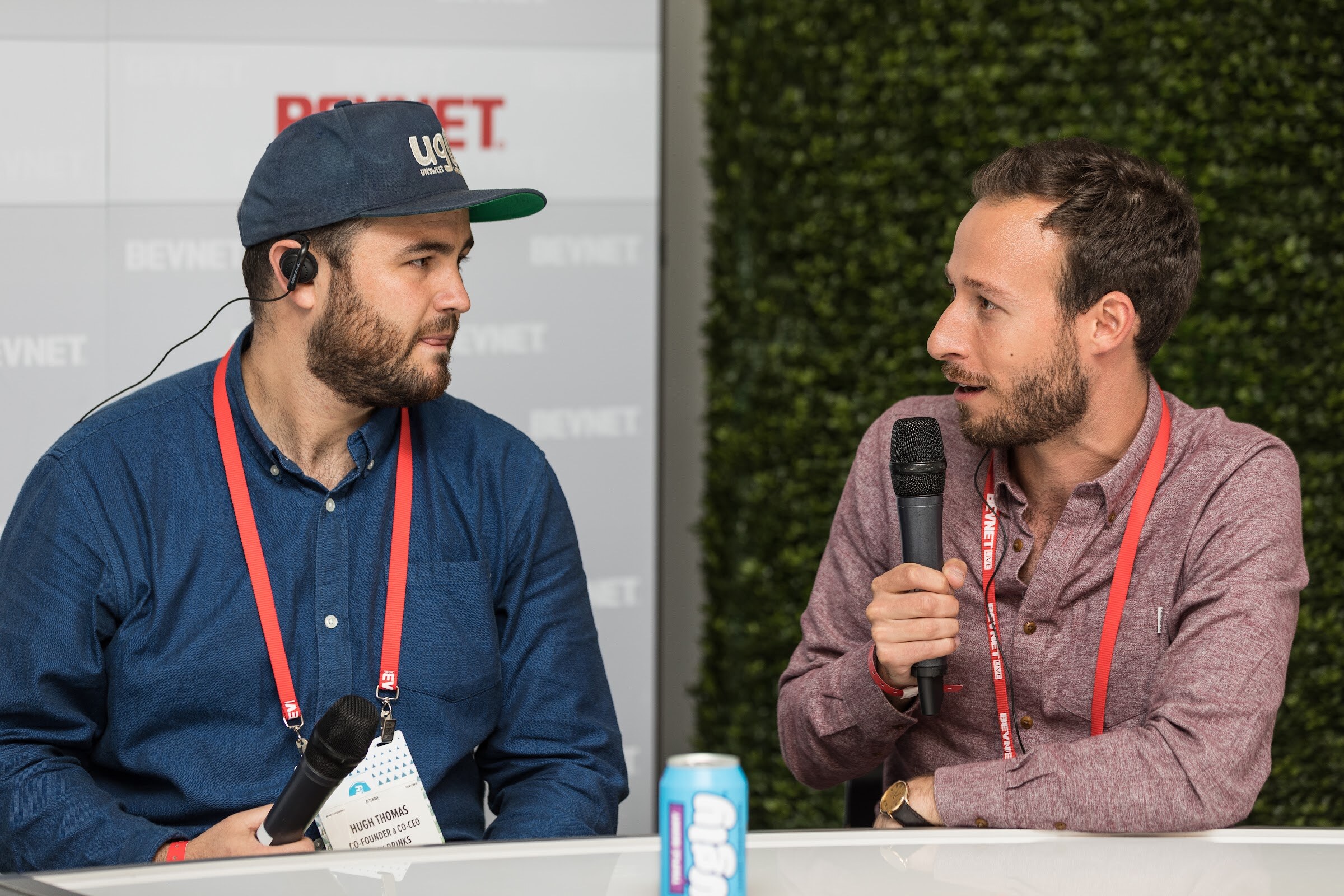 Three men with handheld microphones at a white curved desk in front of a BEVNET logo wall; the man at right wears a black cap and red lanyard.