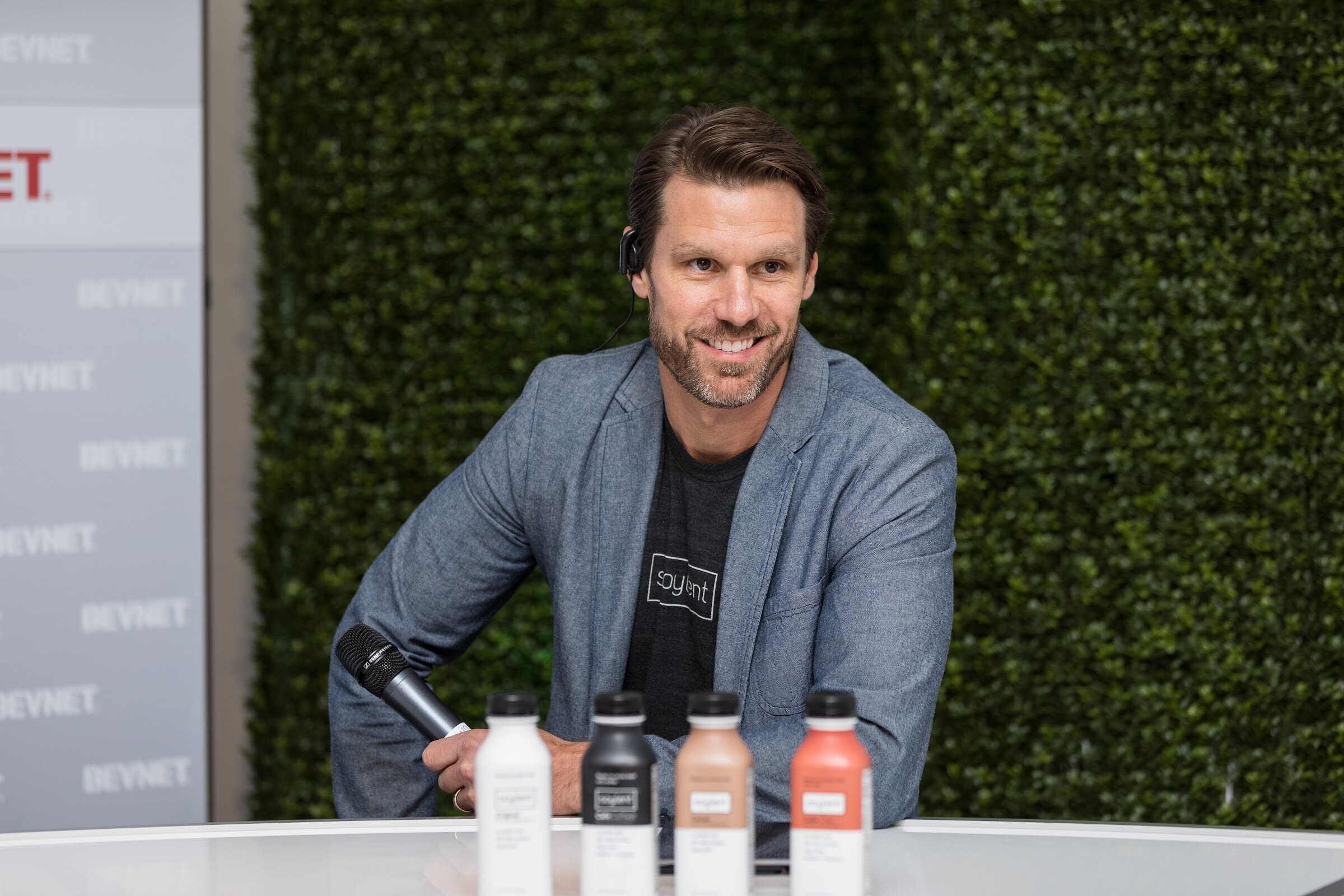 Smiling bearded man in a gray blazer and soylent T-shirt holding a microphone behind four bottles of Soylent on a white desk, with a green hedge wall behind him.