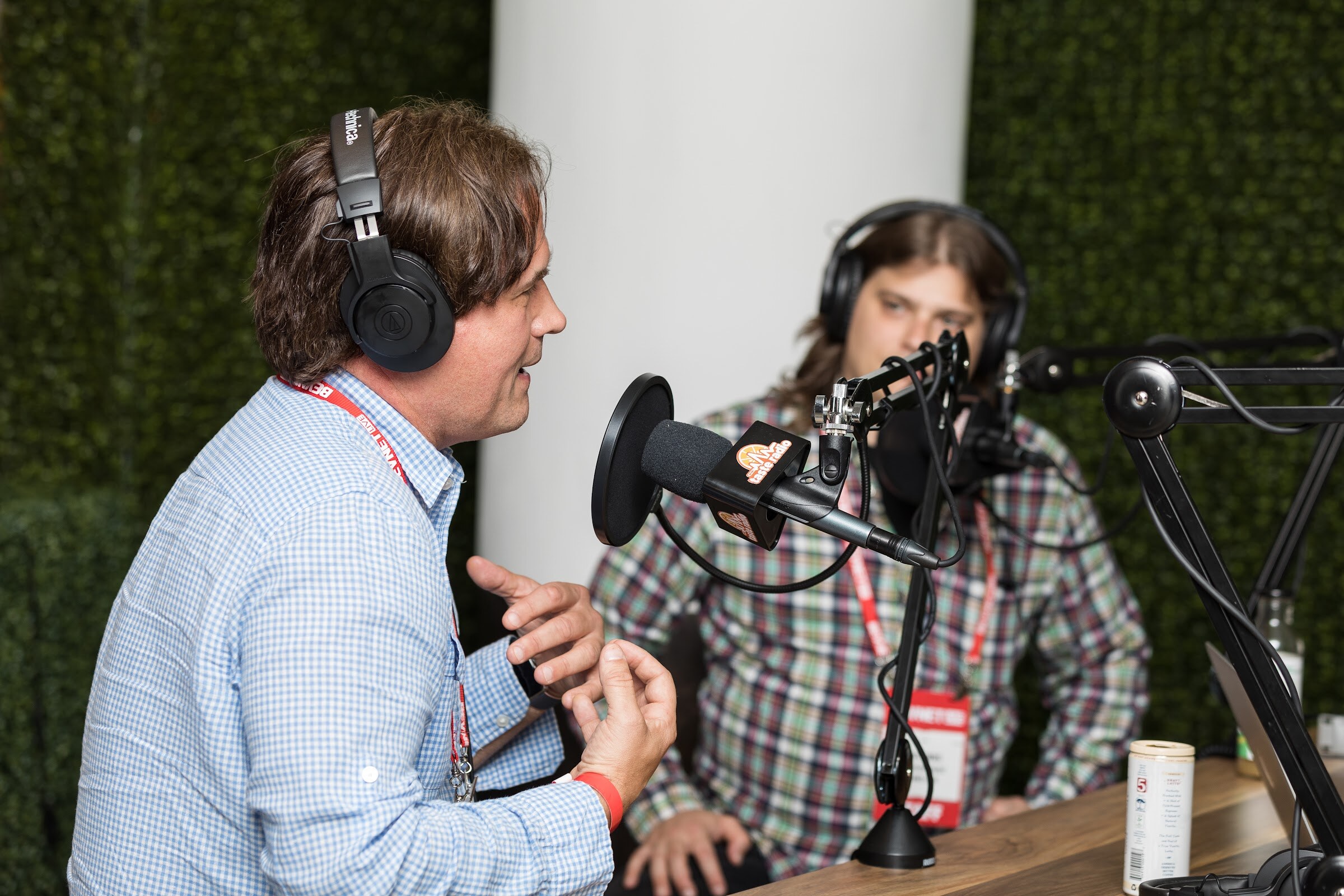 Man in a blue gingham shirt and headphones speaking while a person in a plaid shirt and headphones listens, with a boom-arm microphone and a can on a wooden desk.