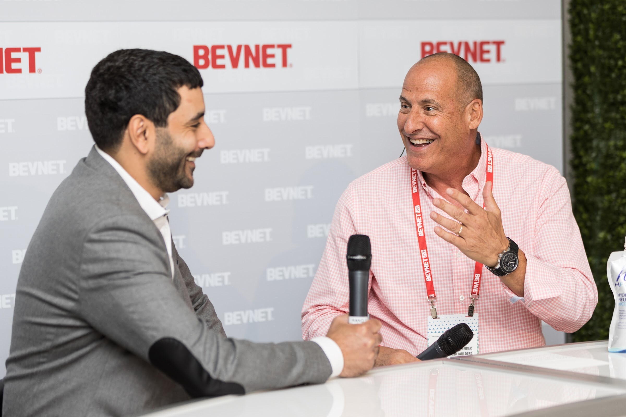 Bald smiling man in a pink checked shirt and BevNET lanyard talking with a man in a gray blazer holding a microphone, in front of a BEVNET wall.