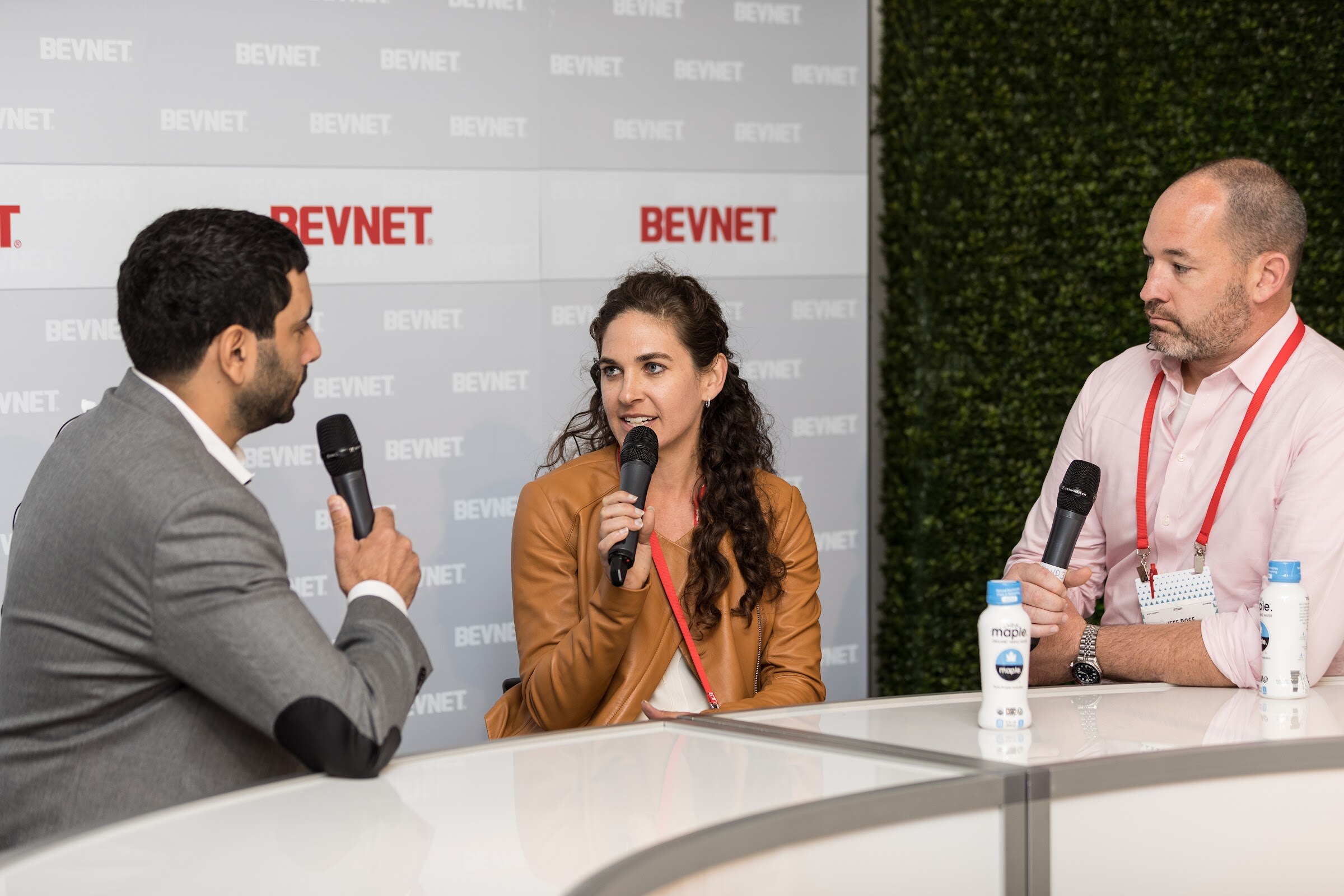 A man in a gray blazer holding a microphone speaks with a woman in a tan jacket and a man in a pink shirt at a white desk with two bottles of maple water.