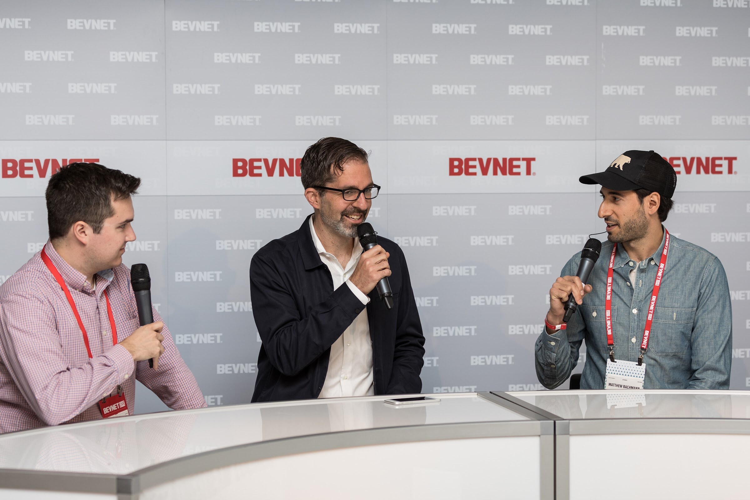 Three men with handheld microphones seated at a white desk in front of a BEVNET logo wall; the man at right wears a black cap and a red lanyard.