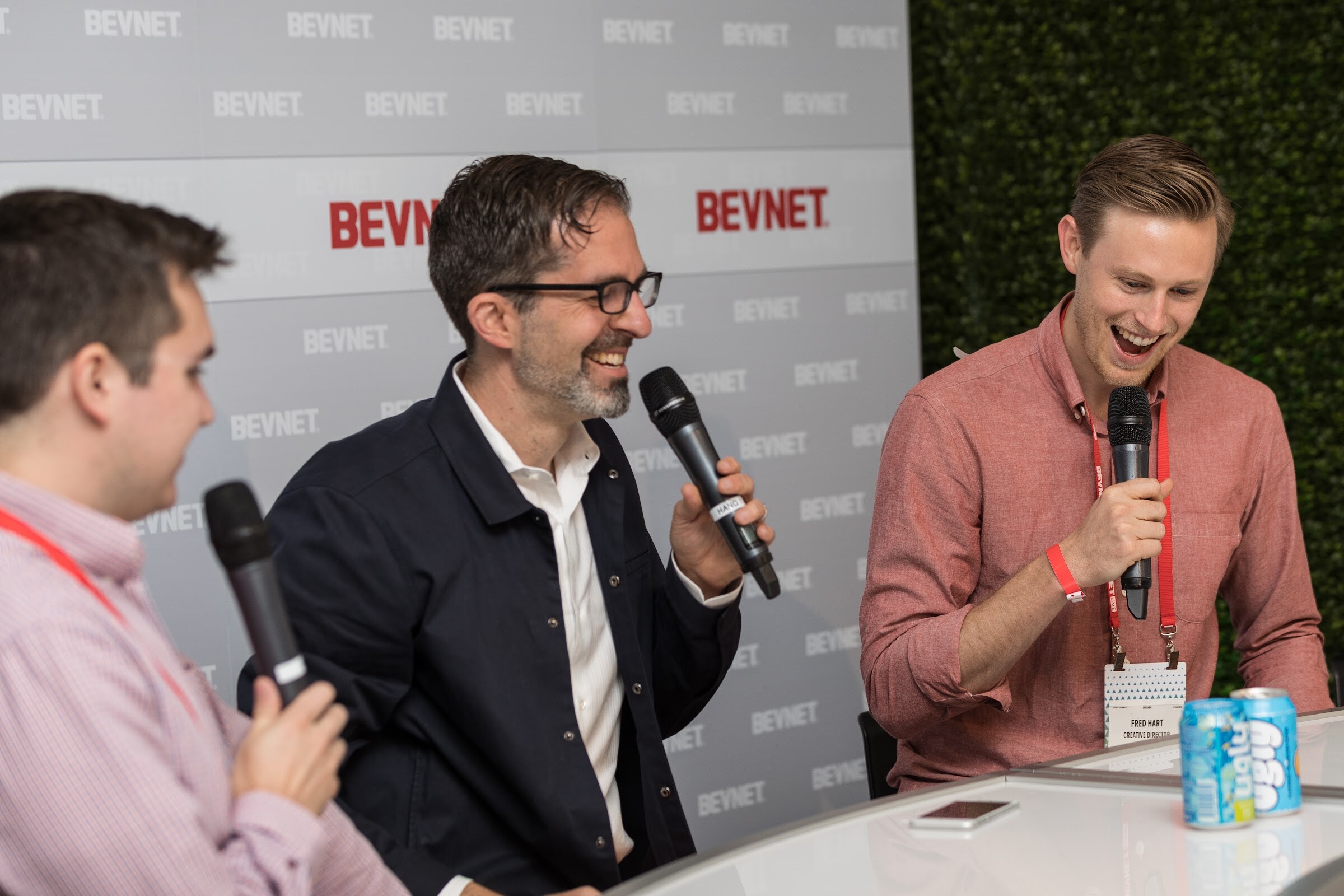 Man in glasses and a navy shirt-jacket speaking into a microphone beside a laughing man in a coral shirt, with two cans on the desk and a green hedge wall behind.