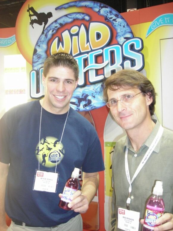 Two smiling men wearing lanyards and name badges stand in front of a Wild Waters sign, each holding a bottle of purple Wild Waters drink.