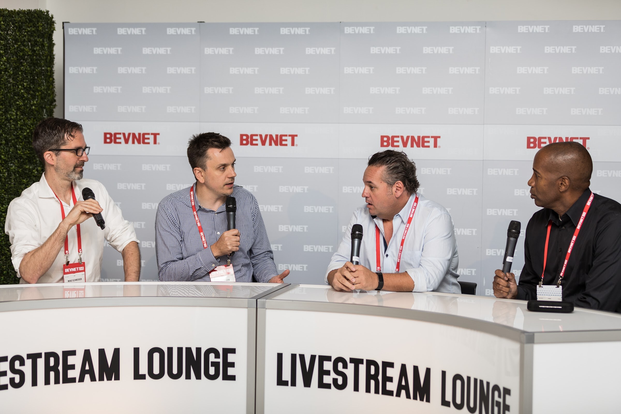 Four men with handheld microphones seated at a white desk marked LIVESTREAM LOUNGE in front of a BEVNET logo wall.