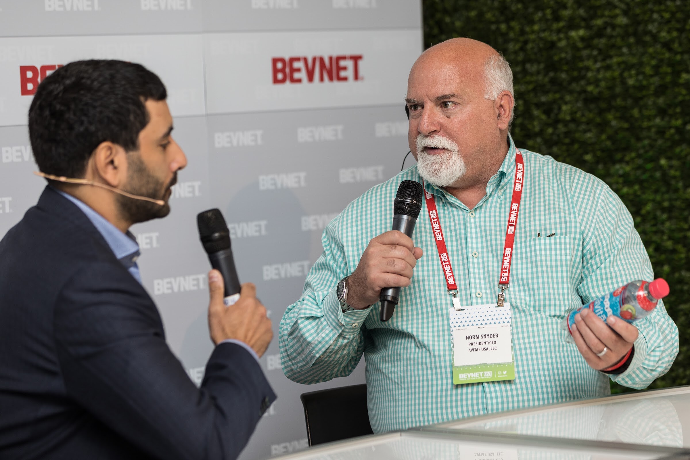 Man with a white beard in a green and white checked shirt, lanyard and name badge, holding a microphone and a bottle of water, speaking with a host in a navy blazer.