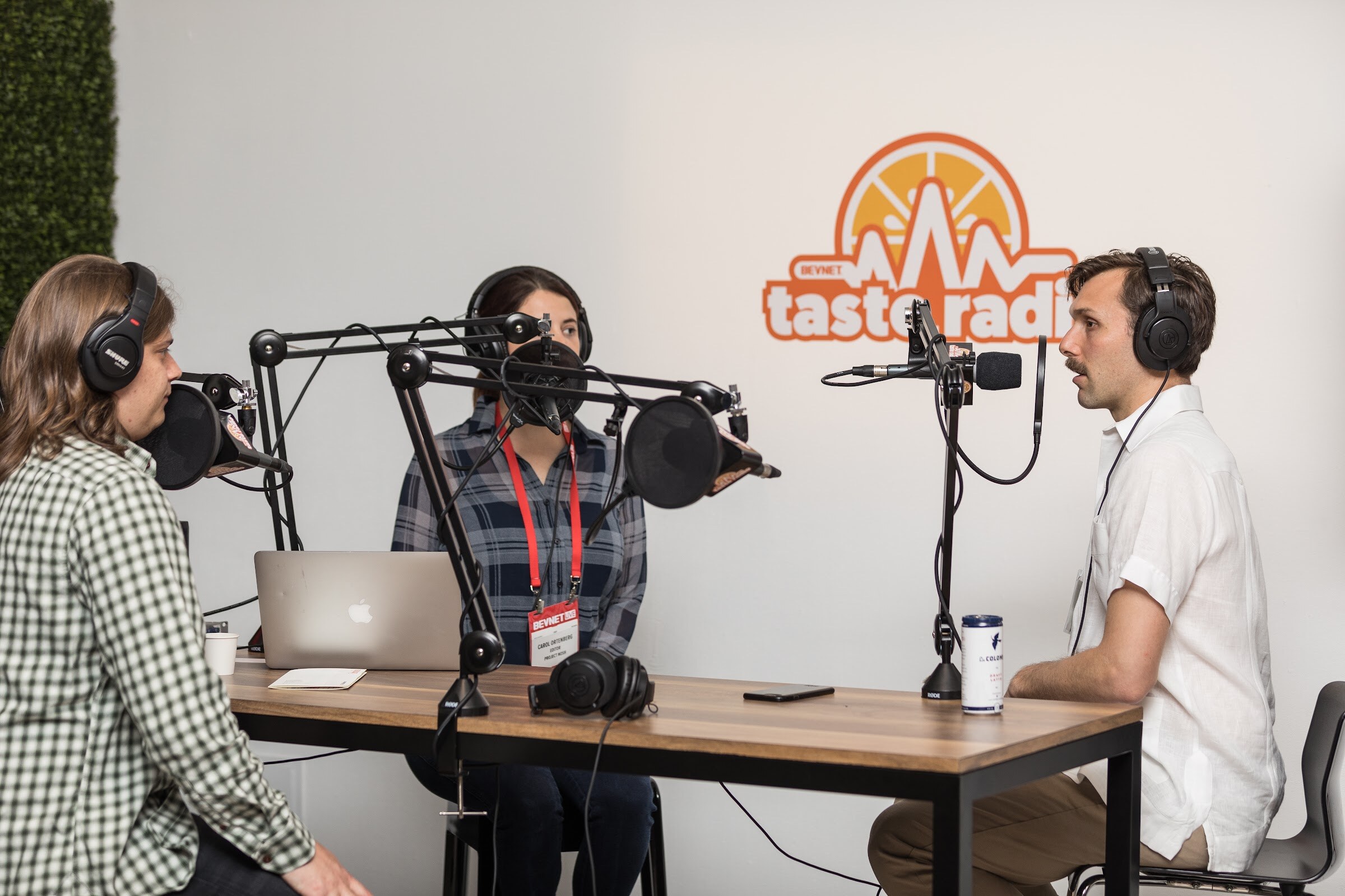 Three people in headphones around a wooden desk with boom-arm microphones, a laptop and a can, in front of a Taste Radio logo on a white wall.