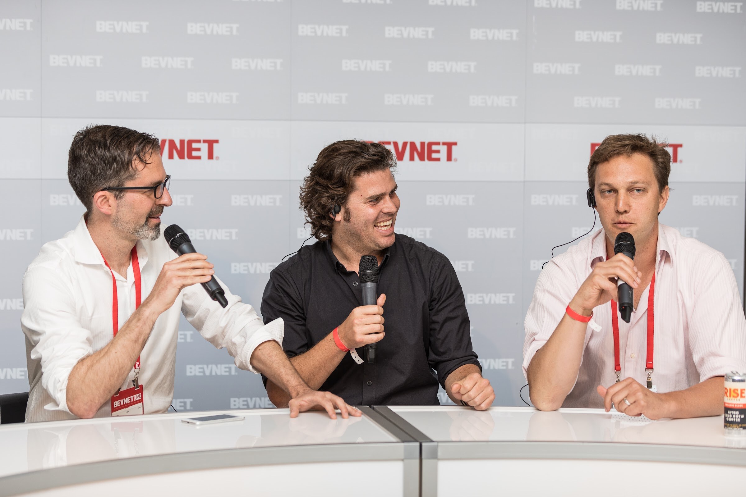 Three men in white and black shirts holding microphones at a white desk in front of a BEVNET wall, one laughing, with a Rise can at the edge of the desk.