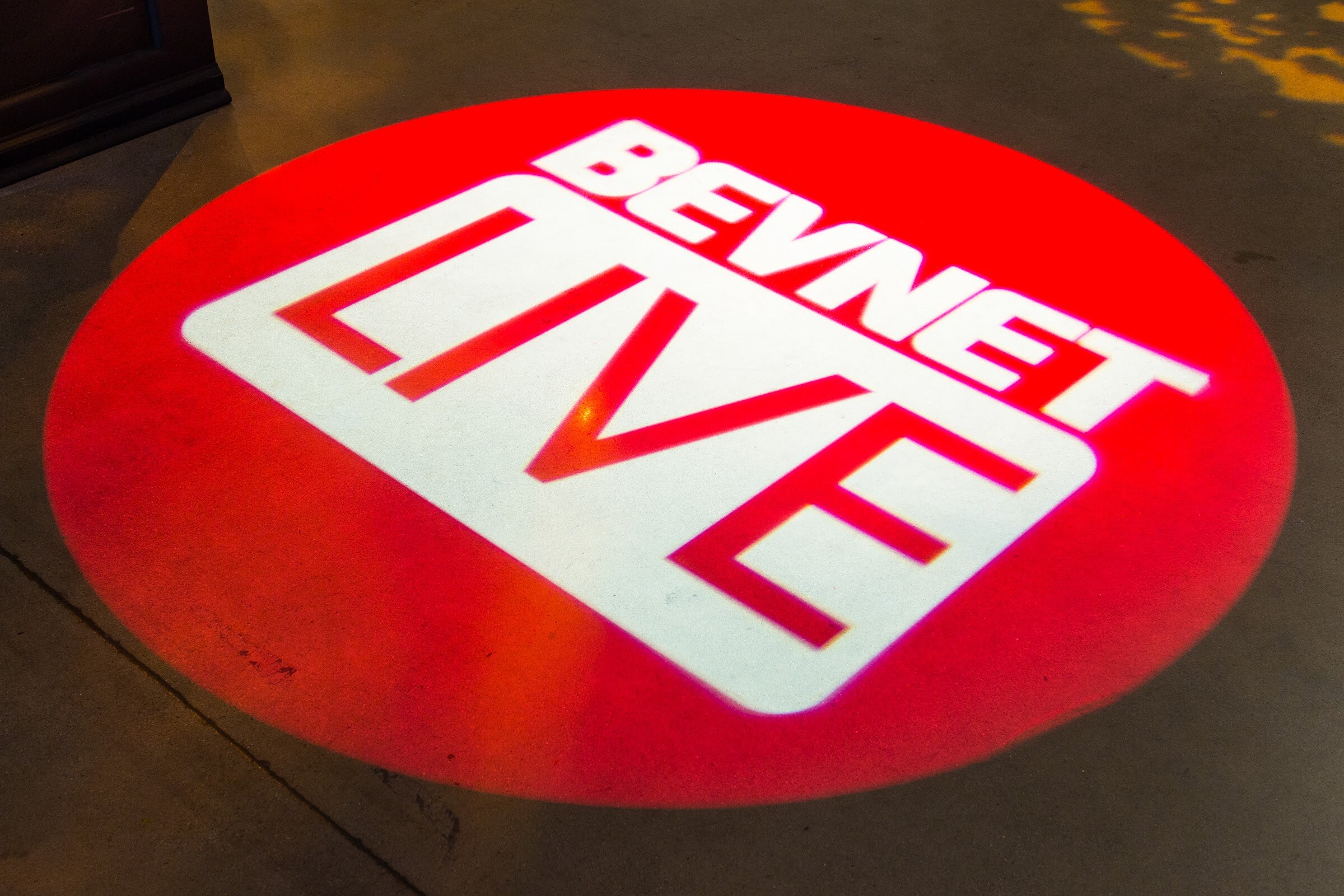Red circular BEVNET LIVE logo projected onto a concrete floor.