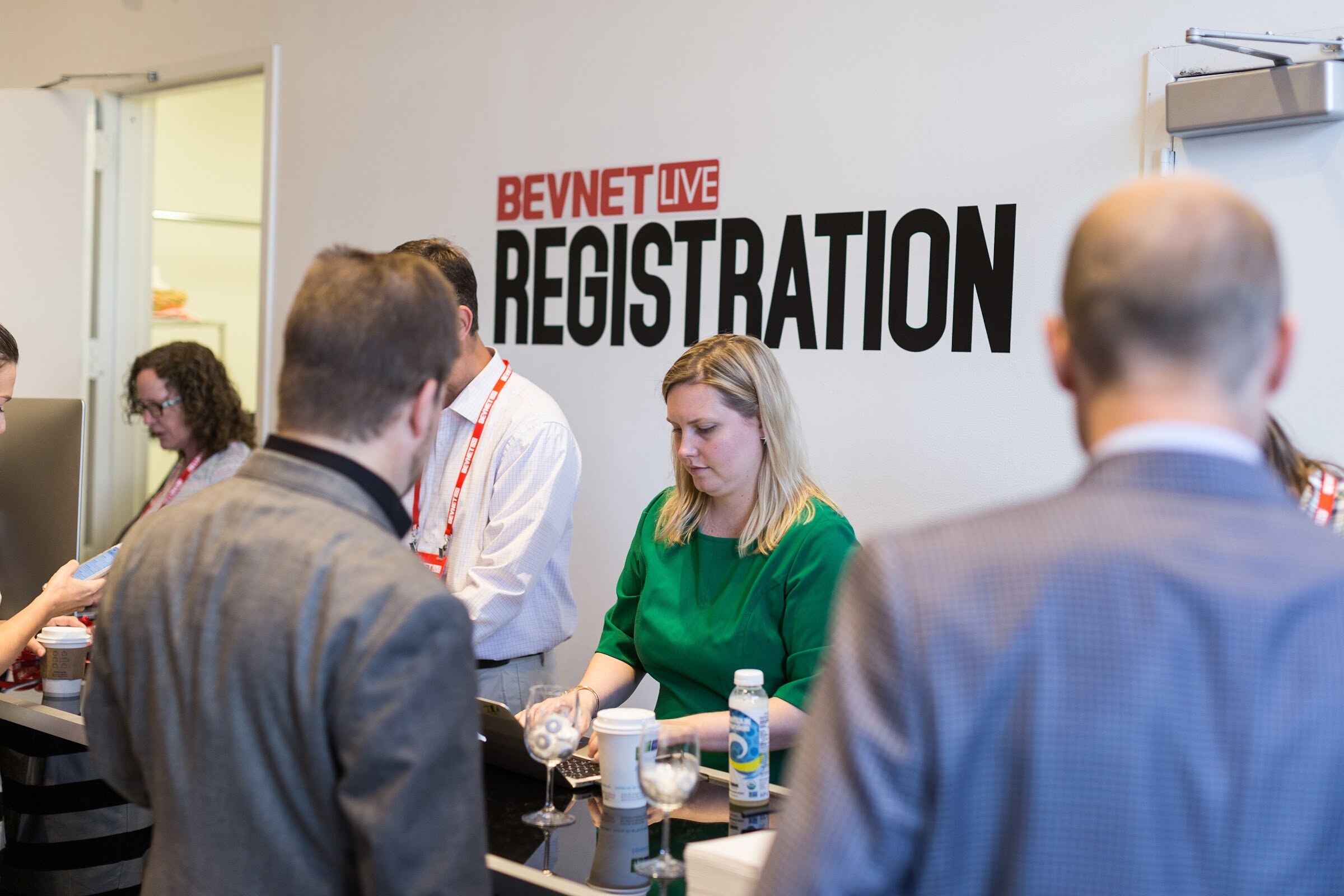 Woman in a green top at a registration counter under a BEVNET LIVE REGISTRATION wall sign, with attendees in jackets in the foreground.
